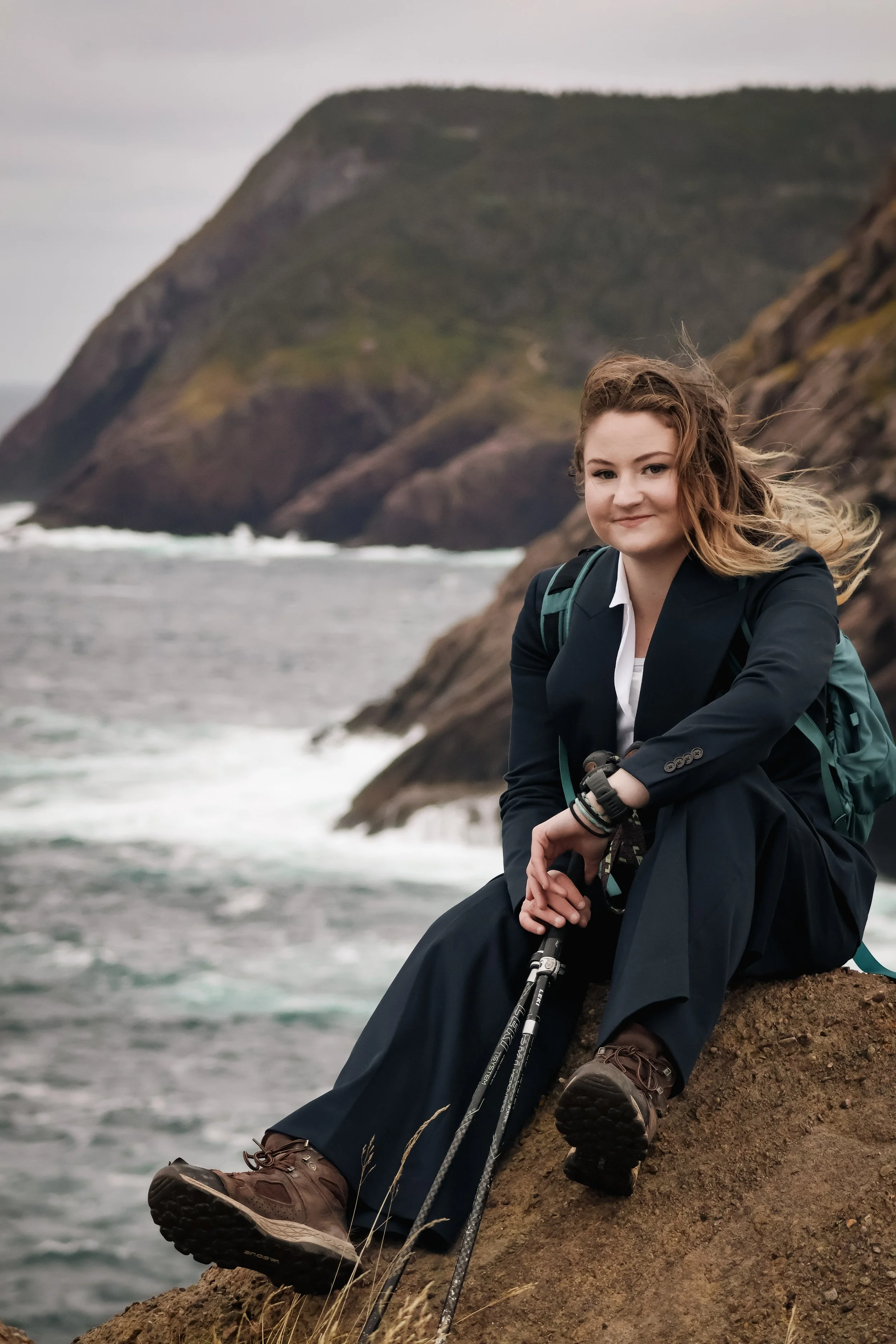 A woman in outdoor hiking gear sitting on a rocky cliff by the ocean with mountains in the background.