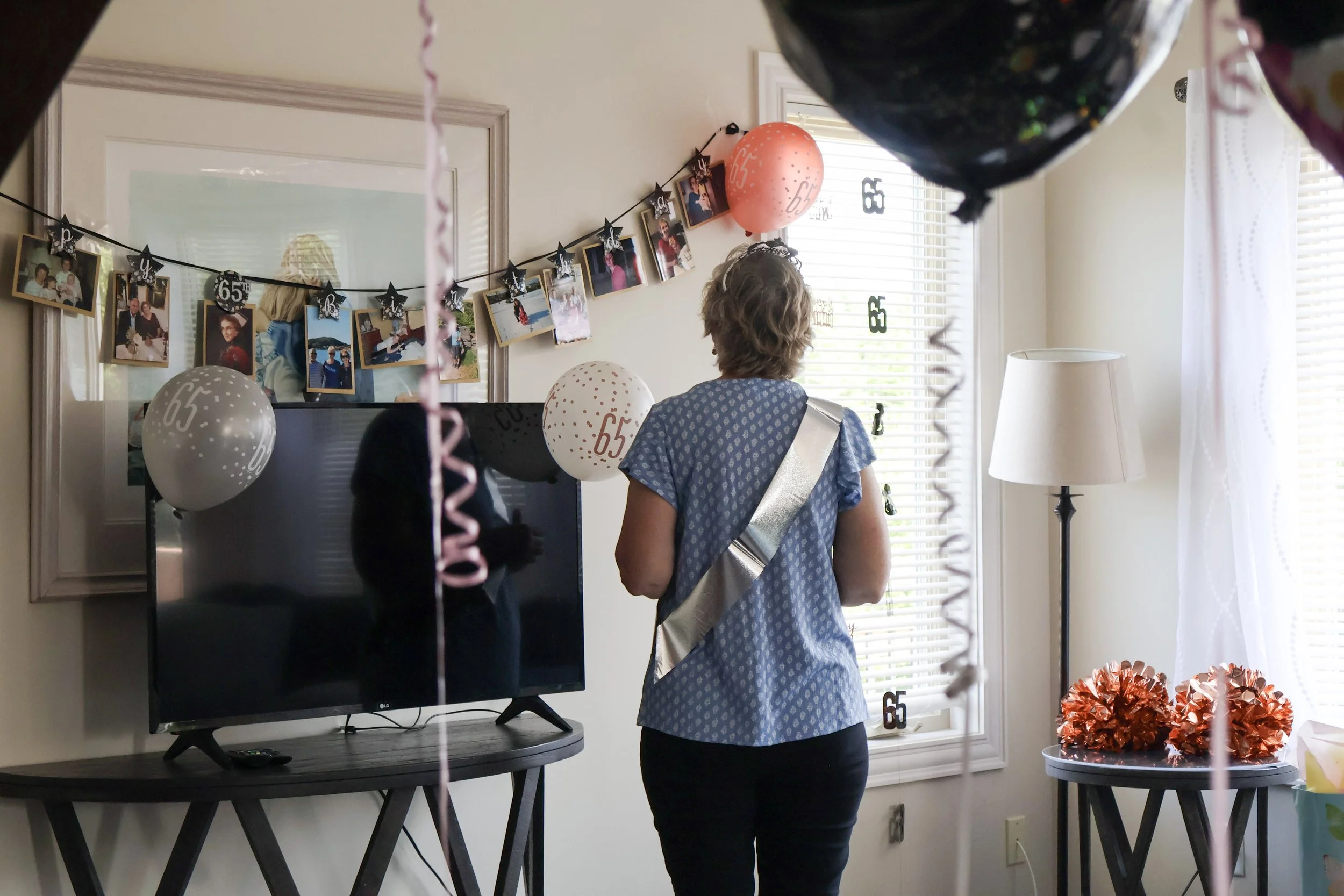 A woman standing in a living room decorated with birthday party decorations, including balloons and photos hanging on a string, celebrating someone's 65th birthday.