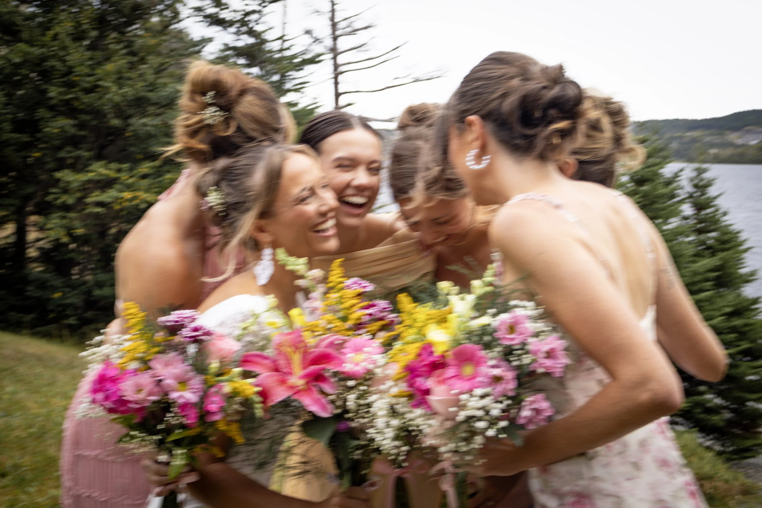 A group of women dressed for a wedding, embracing and laughing outdoors, holding colorful bouquets, with trees and water in the background.