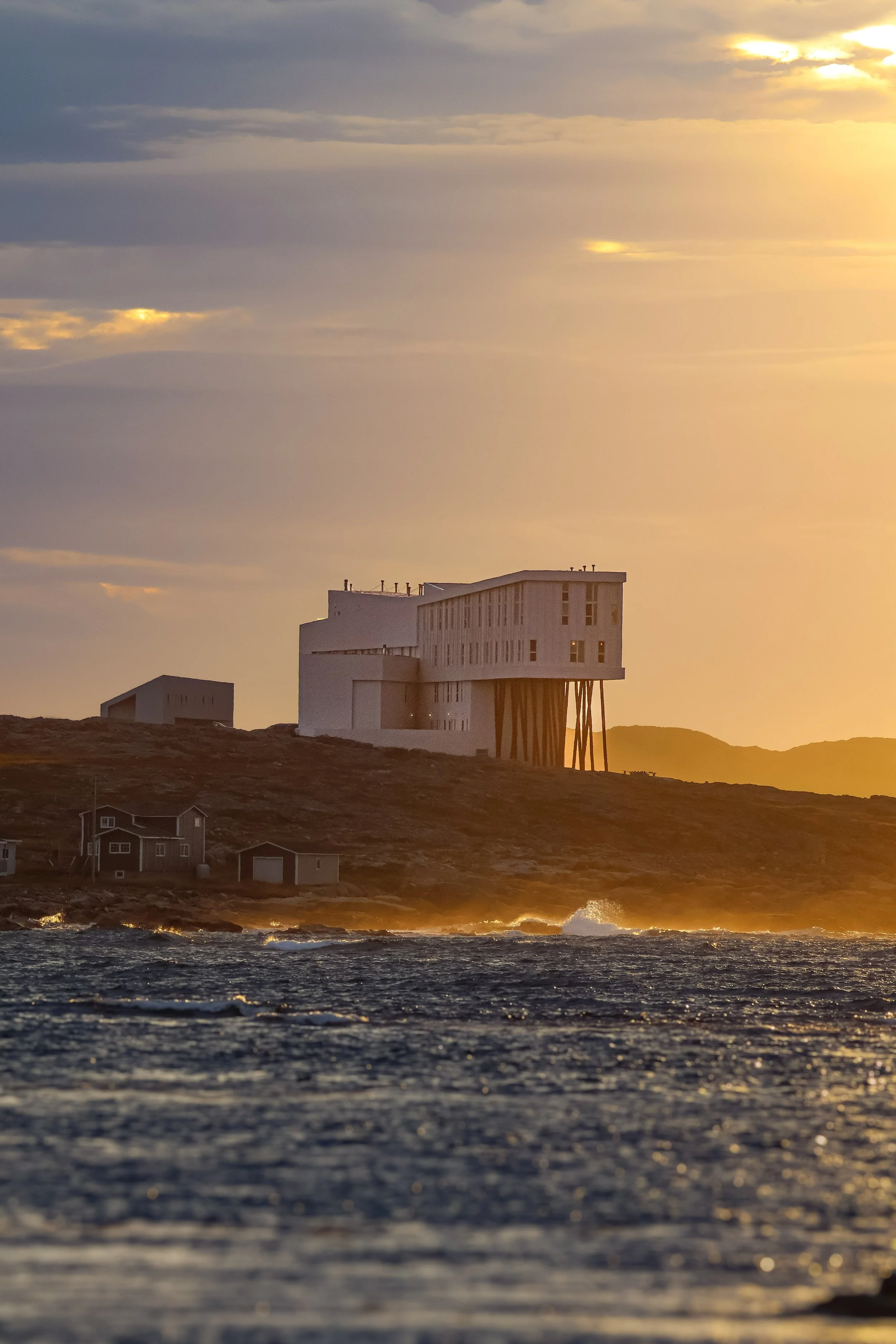 A modern, multi-story house on stilts on a hill overlooking the ocean at sunset, with waves crashing on the shore and a partly cloudy sky.