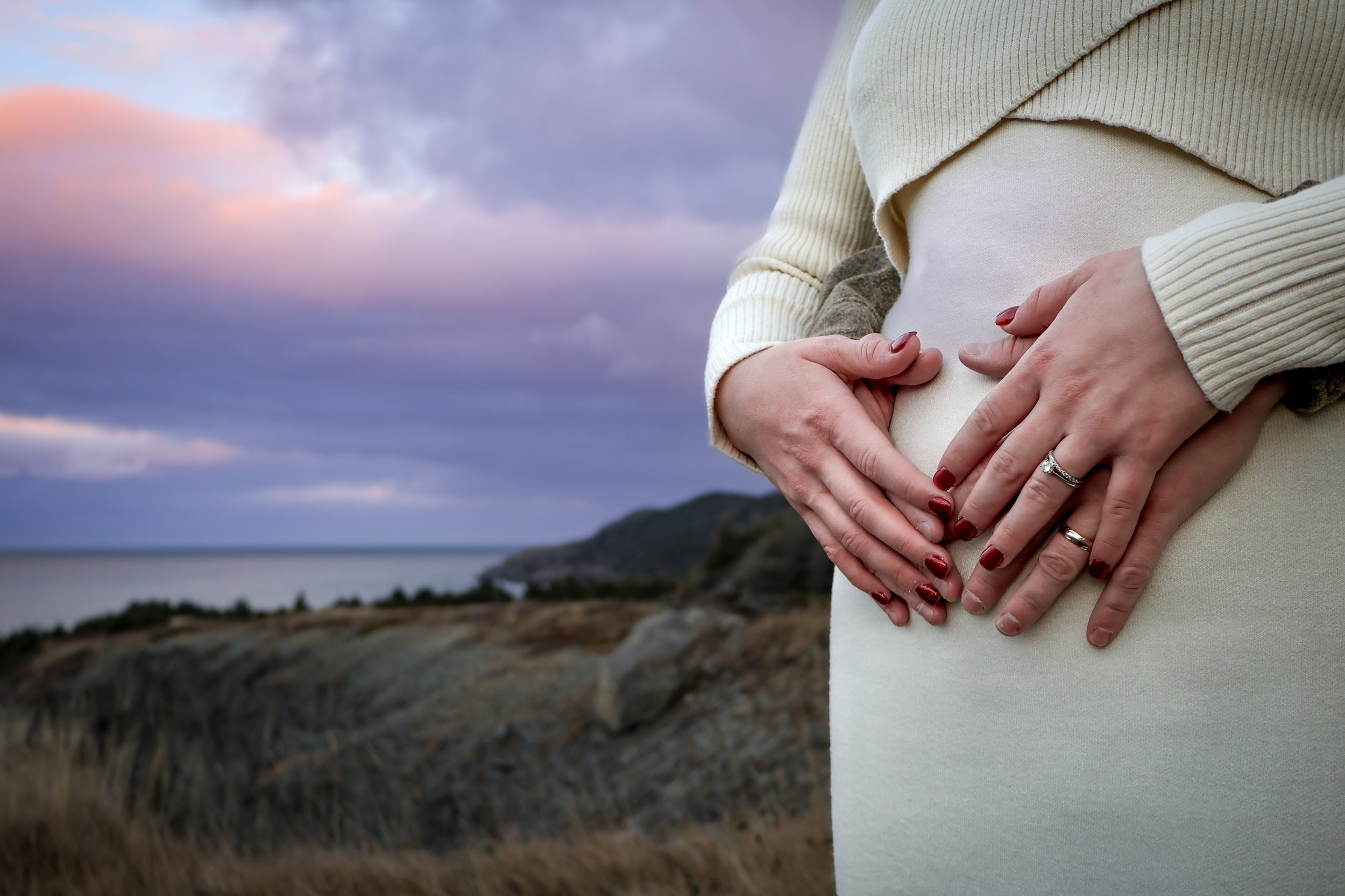 A pregnant woman wearing a cream-colored dress and sweater is holding her belly with both hands, outdoors near a coastline with hills and cloudy sky.