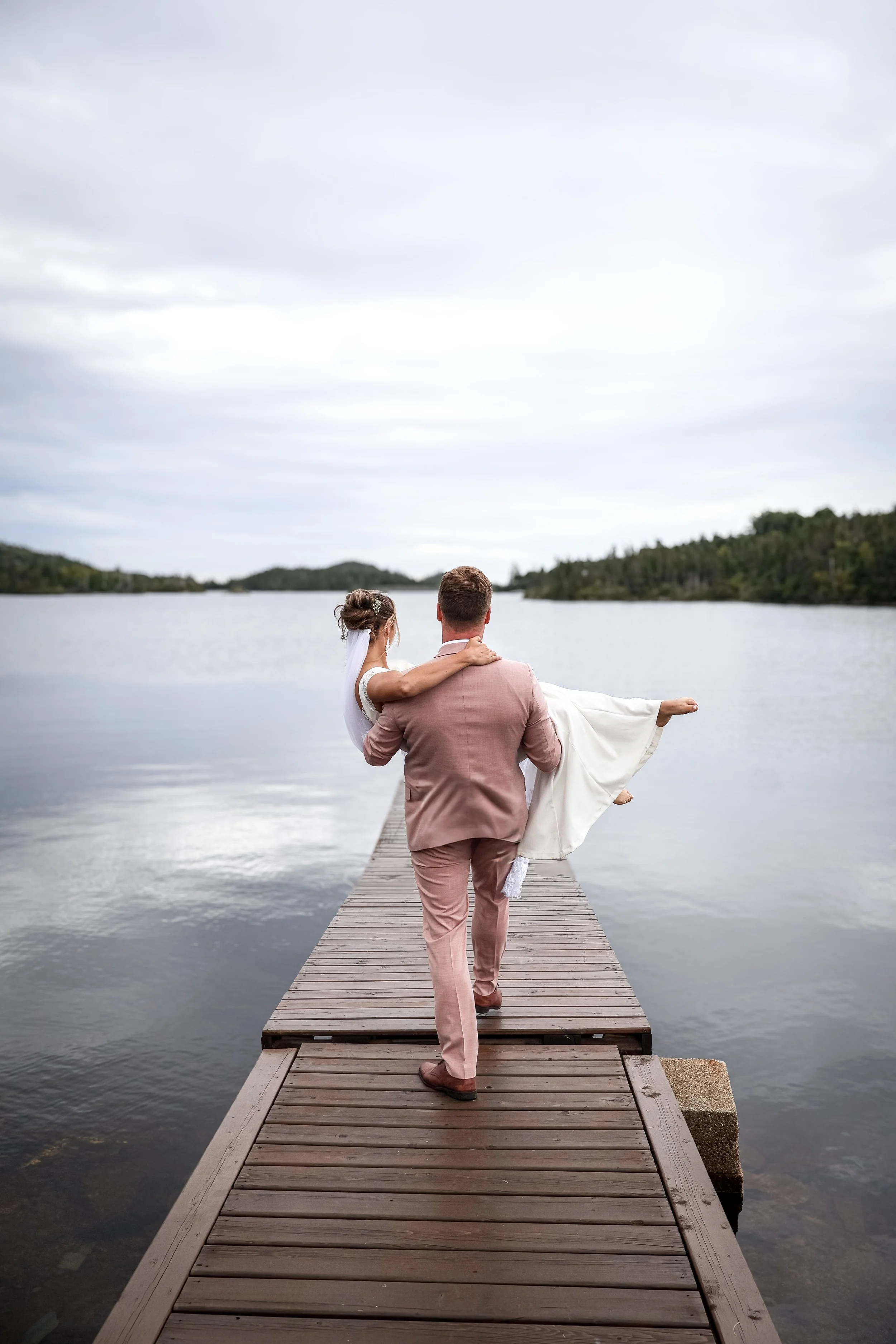 A newlywed couple walking on a wooden dock by a lake, with the groom carrying the bride. The bride is wearing a white wedding dress and veil, and the groom is in a pink suit.