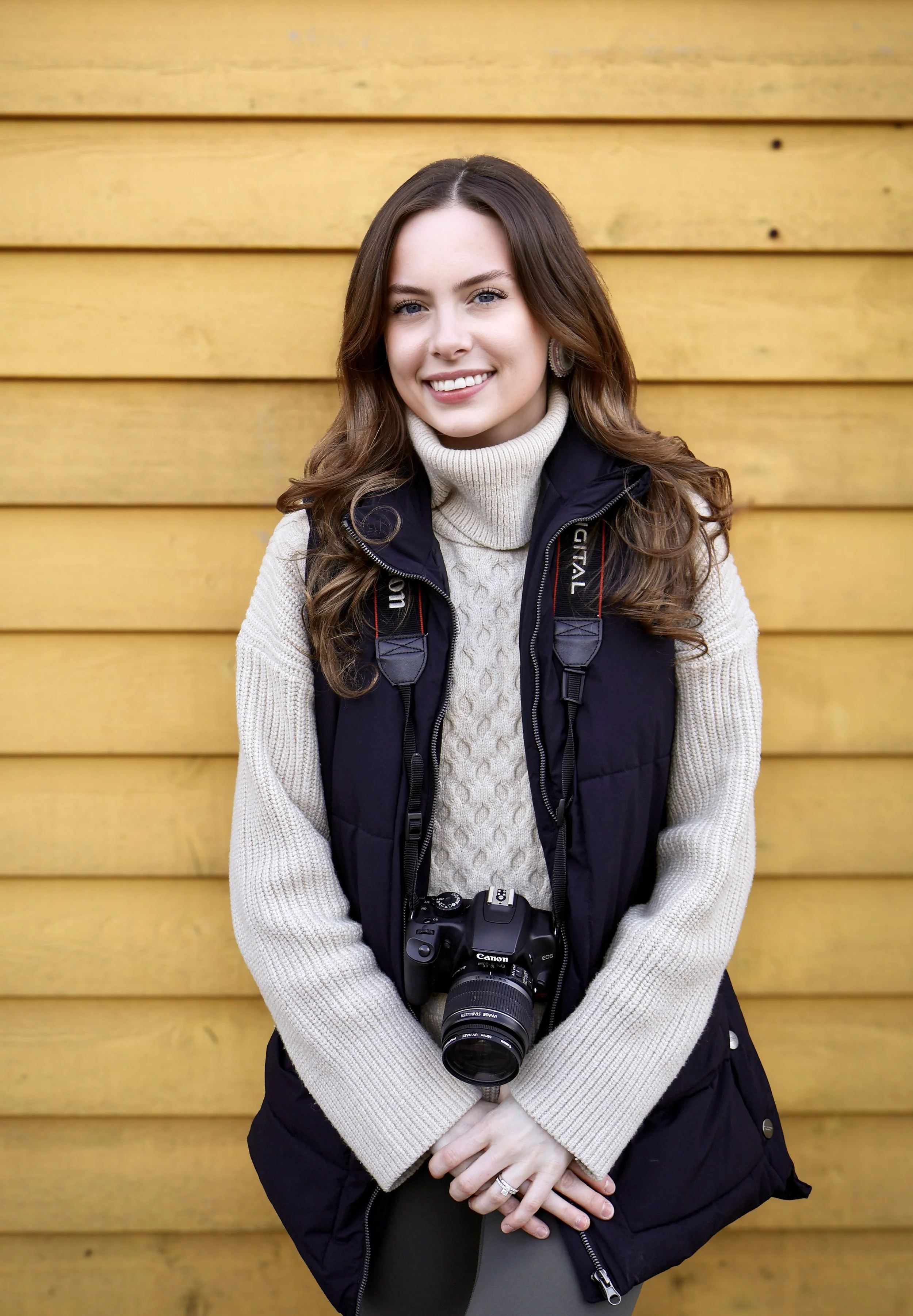 A young woman with long brown hair, wearing a cream turtleneck sweater and a black vest, standing in front of a yellow wooden wall, smiling and holding a camera.