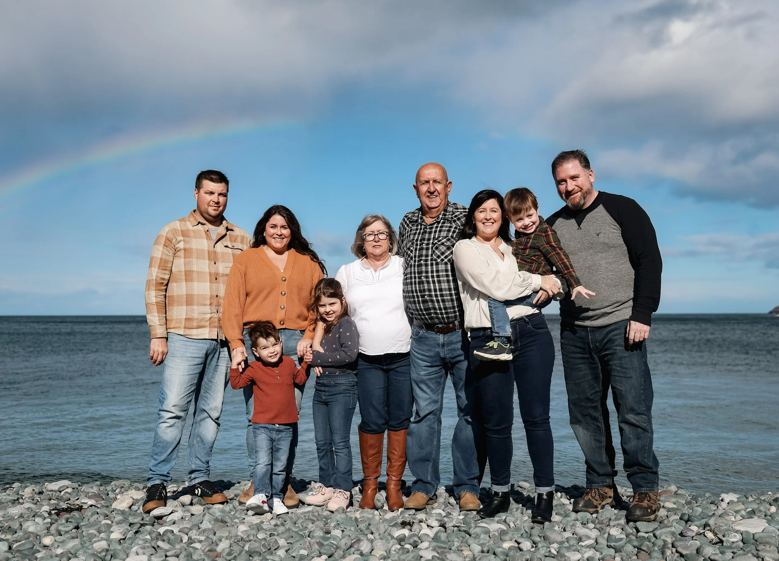 Family of ten standing on a rocky beach with ocean and a rainbow in the sky behind them.