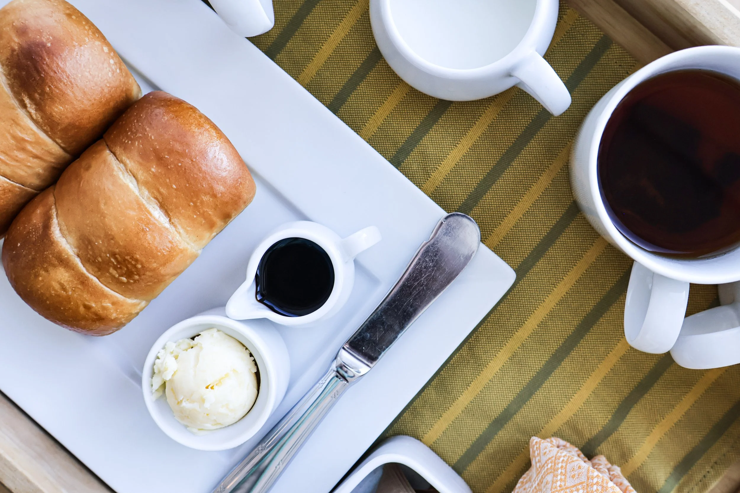 Breakfast tray with two bread rolls, a small cup of butter, a small pitcher of syrup, and a cup of black coffee on a yellow and green striped tablecloth.