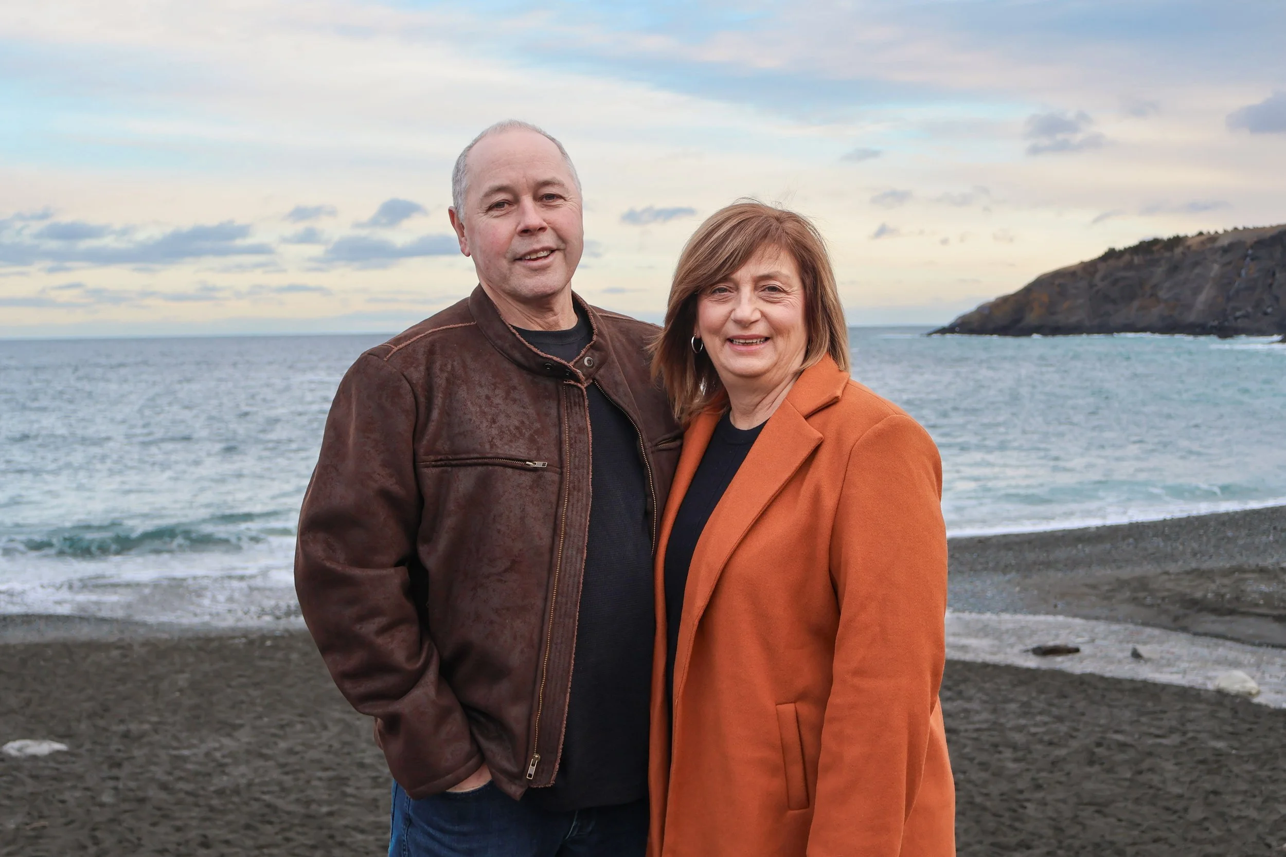 A man and woman standing on a beach near the ocean with hills in the background during sunset or sunrise.