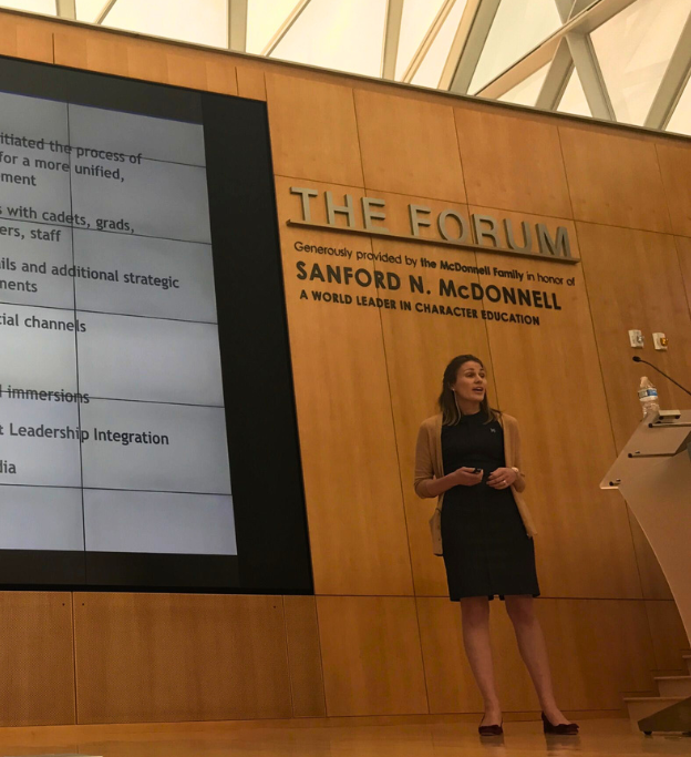 A woman giving a presentation in a conference room called The Forum, with a large screen and a wooden wall behind her.