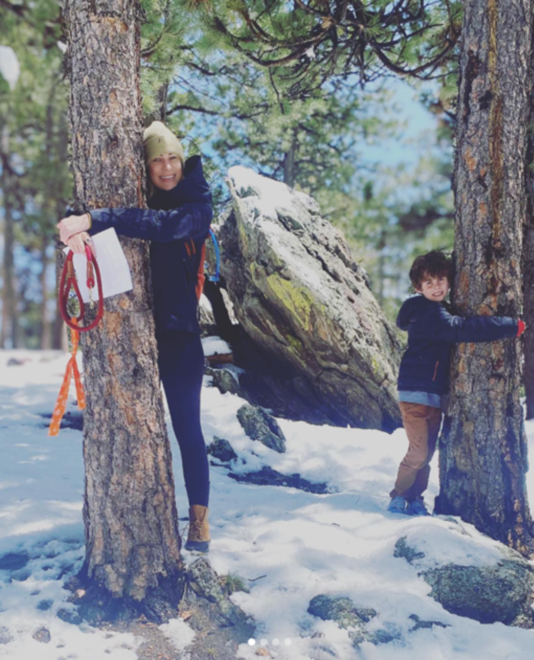 A woman and a young boy hiking in a snowy forest, hugging trees, with rocks and snow on the ground, surrounded by pine trees and a clear sky.