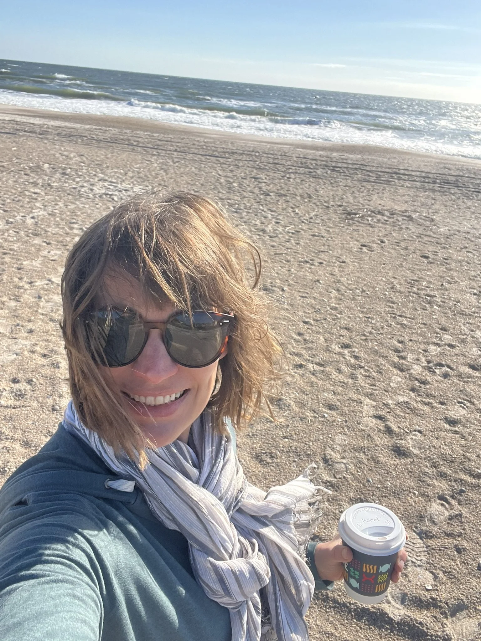 A woman with wavy hair wearing sunglasses and a scarf takes a selfie on a sandy beach, holding a coffee cup, with the ocean and clear sky in the background.