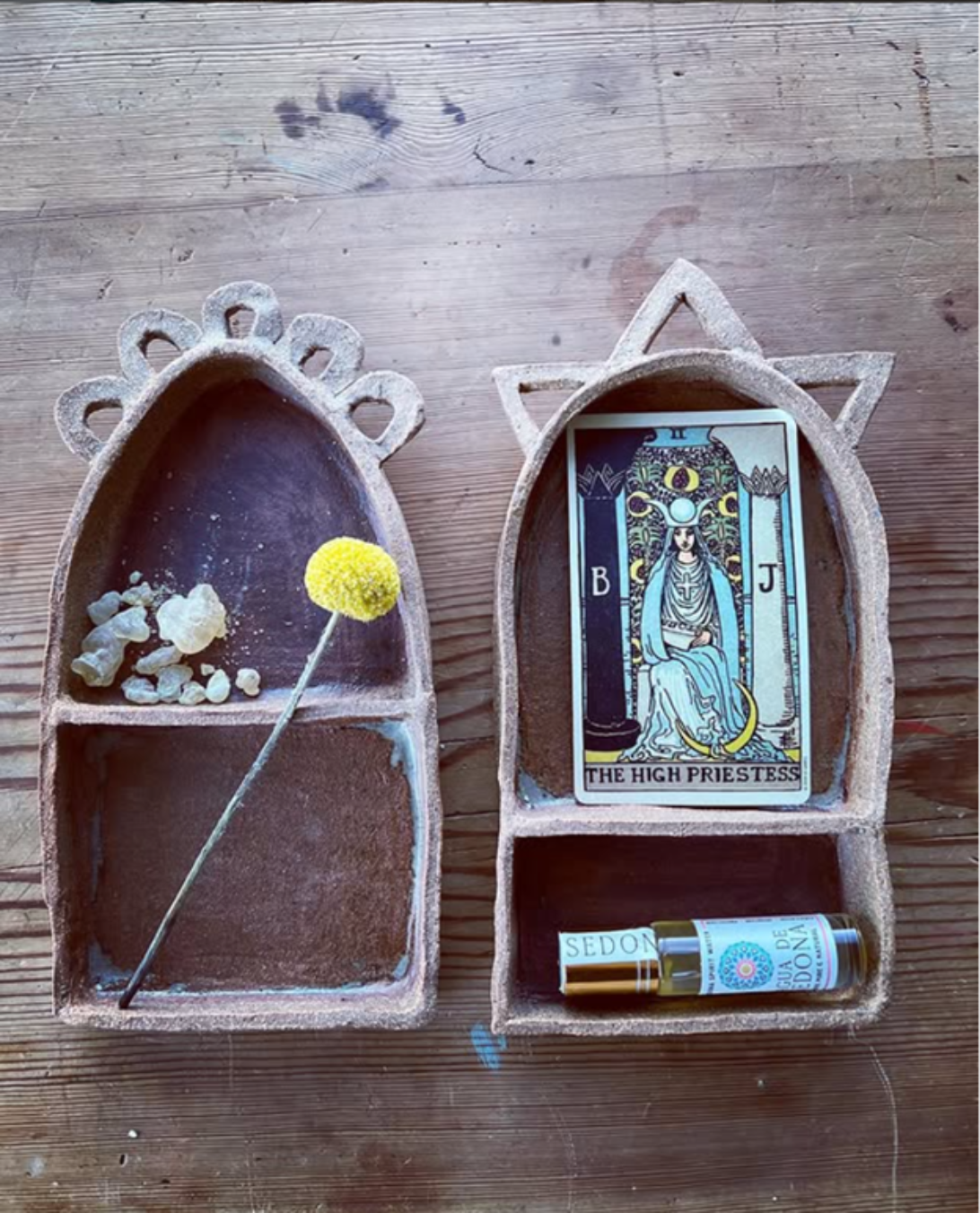 Two small shelves with decorative rowns, one holding a single yellow flower and white stones, the other holding a Tarot card titled 'The High Priestess,' and a small bottle of perfume.