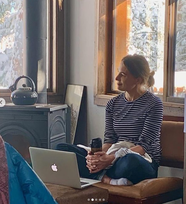 Woman in striped shirt sitting by window holding a mug, with a silver laptop open on her lap.
