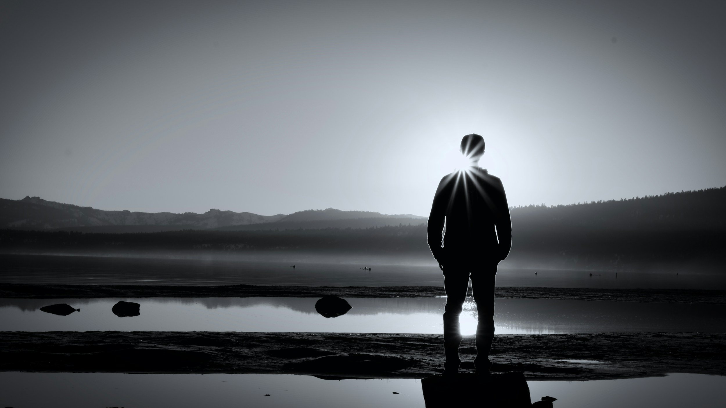 Eine silhouettierte Person steht am Ufer eines Sees mit Bergen im Hintergrund, Sonnenstrahlen hinter dem Kopf und in Schwarz-Weiß-Optik.