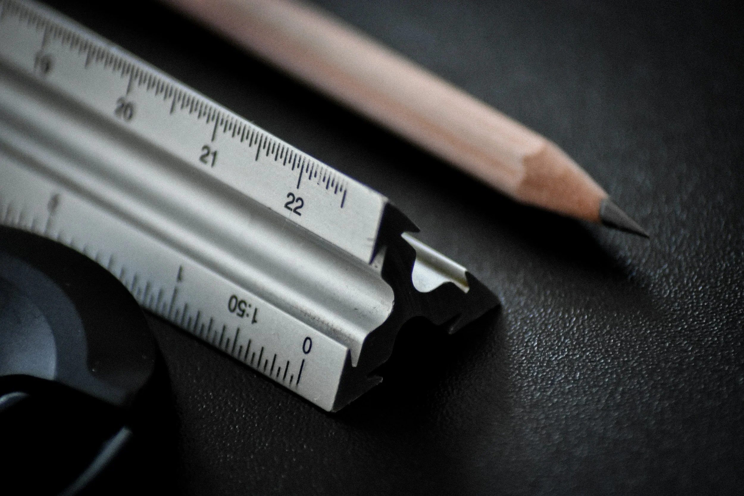 A close-up of a metal retractable ruler with measurement markings, a wooden pencil with a sharpened tip, and a computer mouse on a black textured surface.