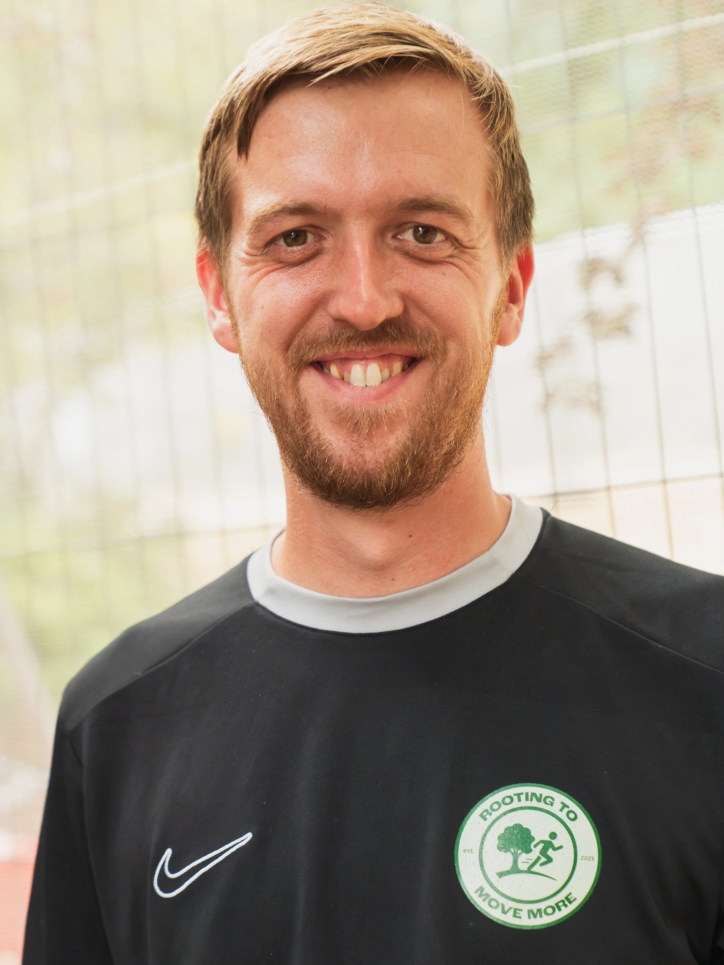 A young man with light brown hair, a beard, and a mustache, smiling and wearing a black athletic jacket with a gray collar, a Nike logo, and a round logo that reads "Rooting to Move More" in blue and green with a running figure and trees.