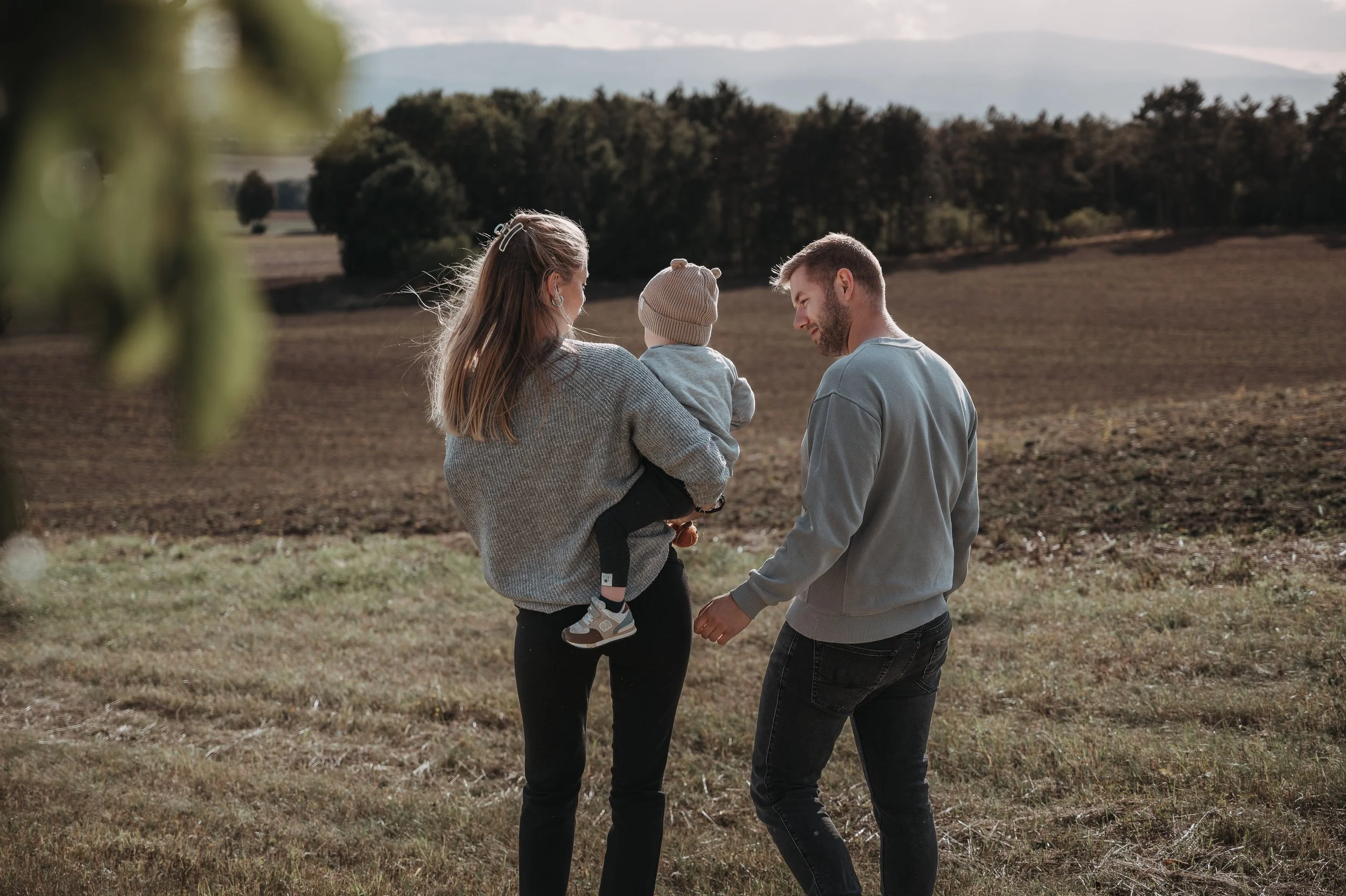 Eine Familie bestehend aus einer Frau, einem Mann und einem kleinen Kind, die in einer ländlichen Umgebung auf einer grünen Wiese spazieren gehen.