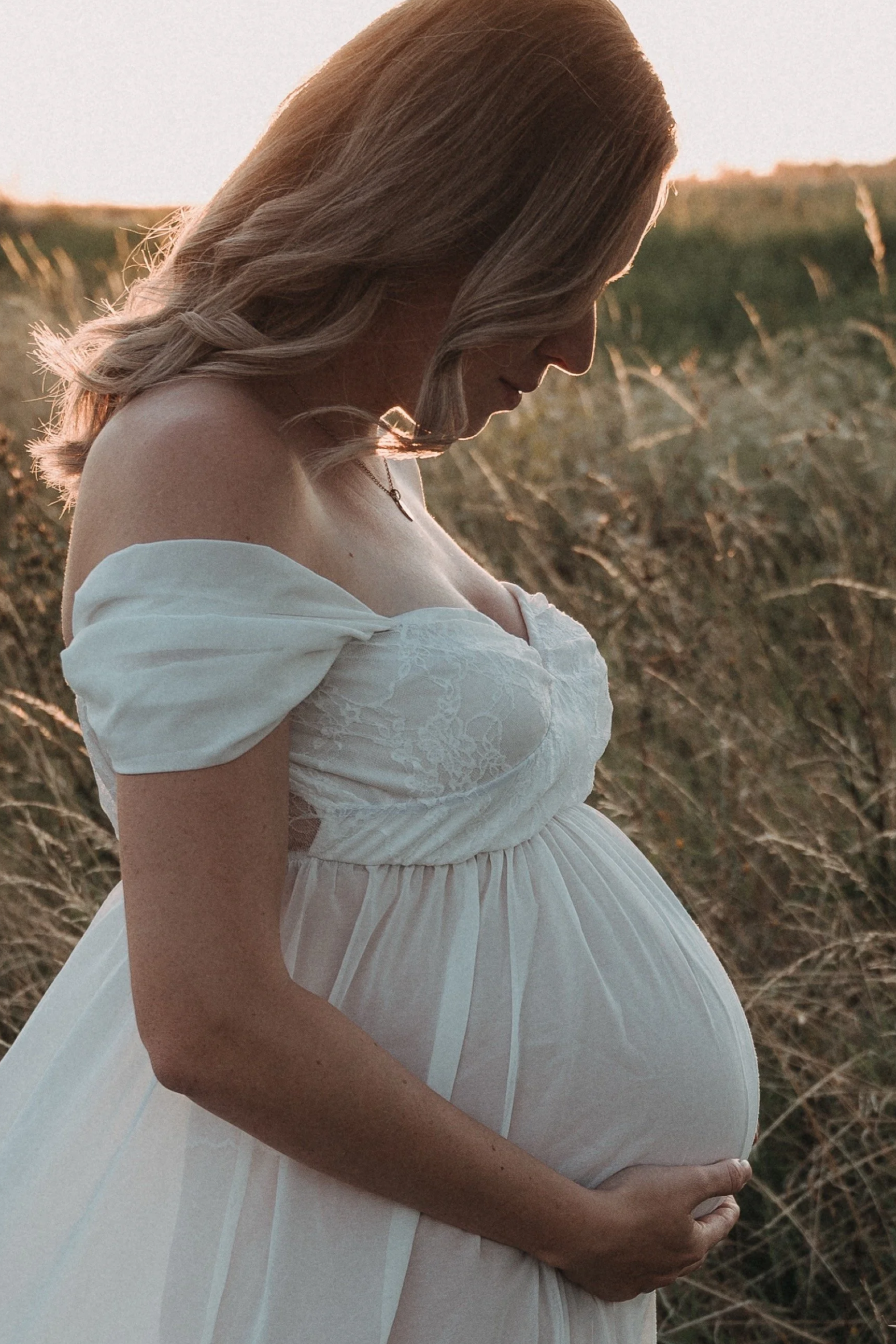 Eine schwangere Frau in einem weißen Kleid steht in einem Feld bei Sonnenuntergang und umarmt ihren Bauch.