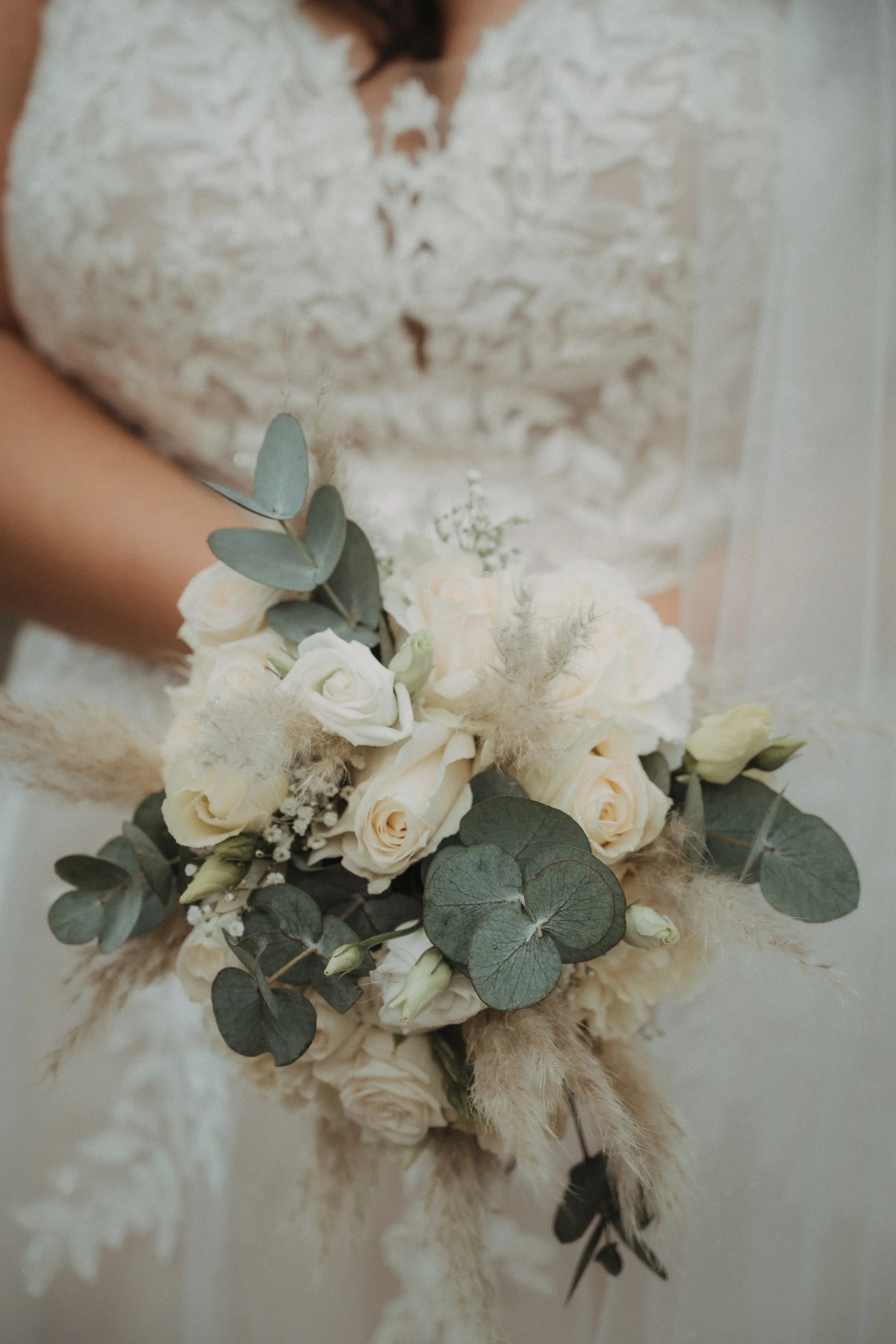 Braut hält einen Brautstrauß mit weißen Rosen, Eukalyptusblättern und getrockneten Pflanzen.