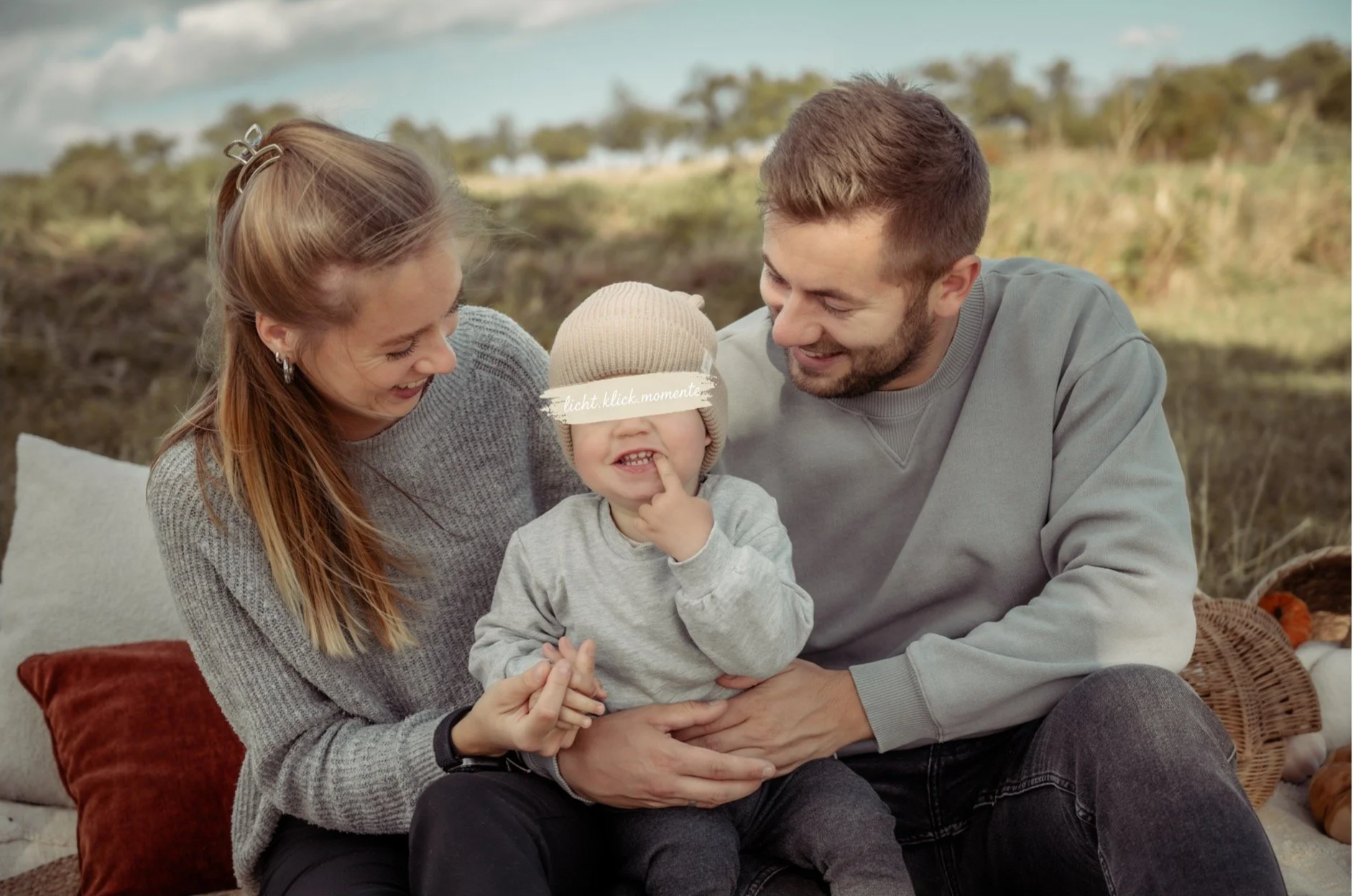 Eine lachende Familie sitzt im Freien auf einem Sofa, bestehend aus einer Mutter, einem Vater und einem kleinen Kind mit Mütze. Das Kind spielt mit seiner Nase, während die Eltern fröhlich zuschauen.