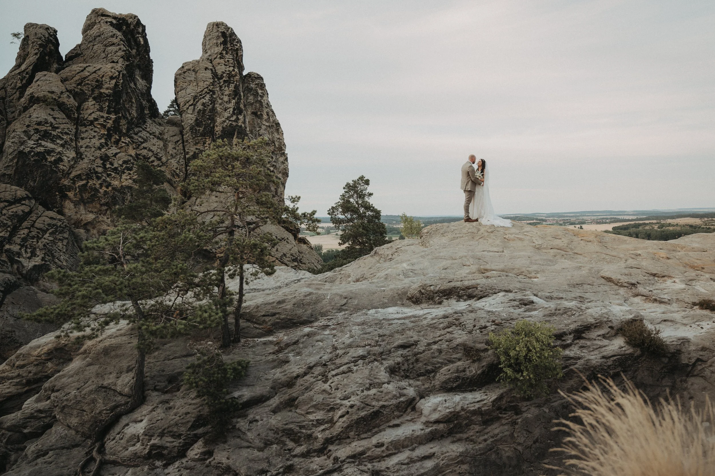 Ein Brautpaar steht auf einem Felsen in einer natürlichen Landschaft, umgeben von Bäumen und großen Felsformationen, mit einer weiten Ebene im Hintergrund.