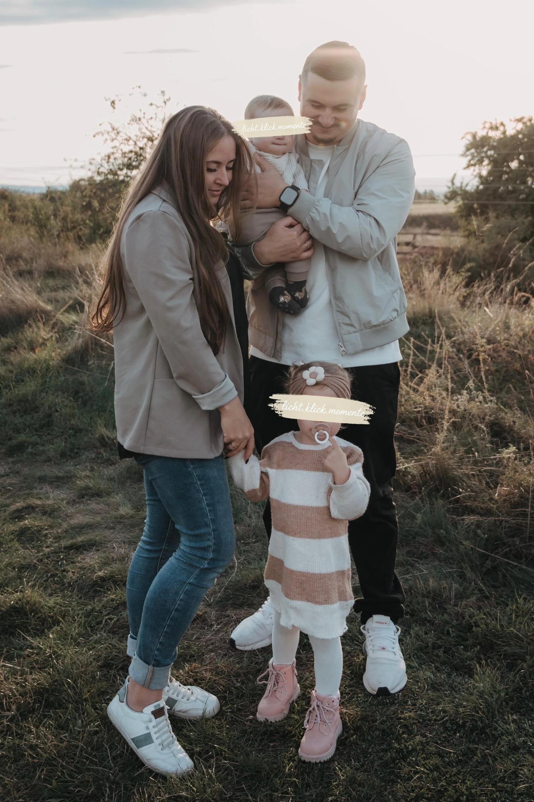 Familie im Freien bei Sonnenuntergang, bestehend aus einem Mann, einer Frau, zwei Kleinkindern und einem Baby, alle lächelnd zusammen. Der Hintergrund zeigt Natur mit Büschen und einem Feld.