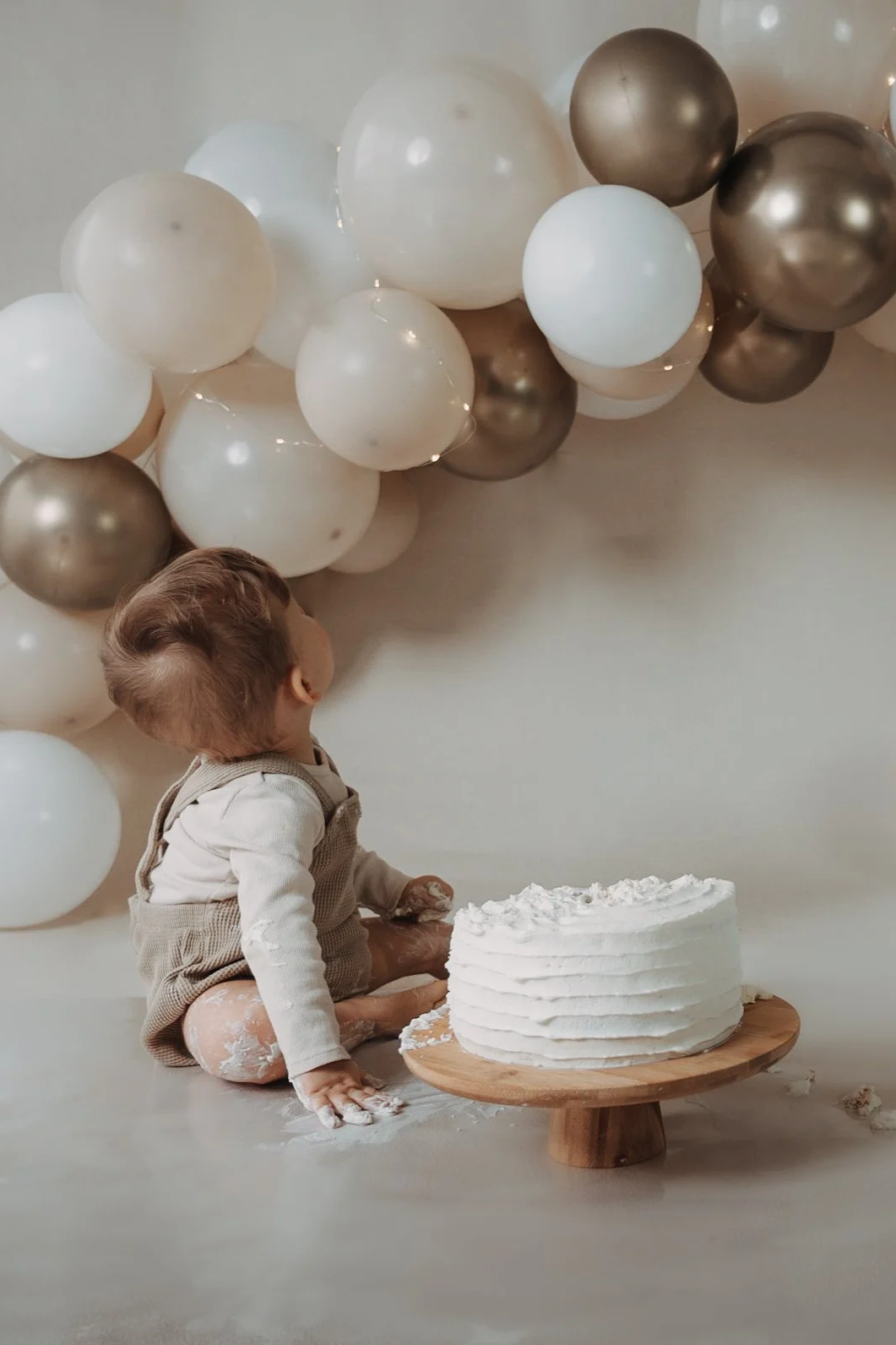 Ein Kind sitzt vor einer weißen Torte auf einem Holzständer, umgeben von weißen und goldfarbenen Ballons, vermutlich bei einer Geburtstagsfeier.