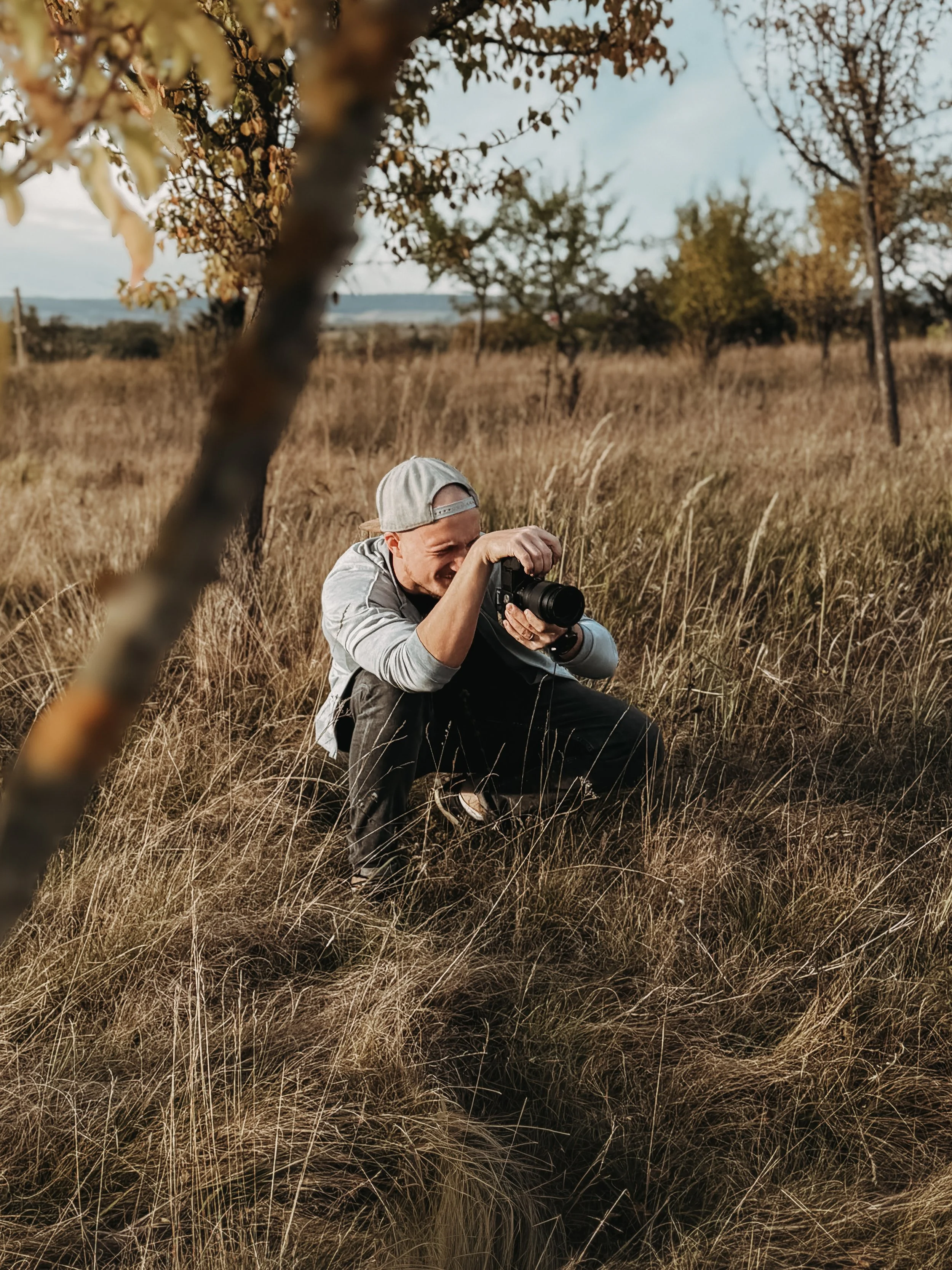Ein Junge im Gras versteckt, fotografiert mit einer Kamera im Freien auf einer Wiese, umgeben von Bäumen und Himmel im Hintergrund.