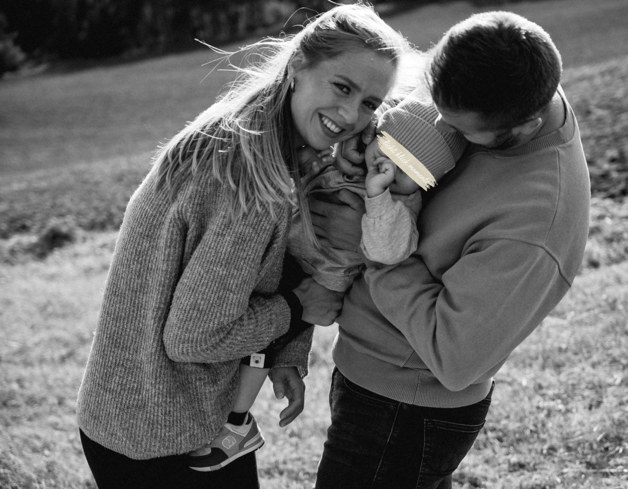 Ein lachendes Paar hält ein Baby in der Natur im Freien. Das Baby trägt einen Pullover mit einem Fernseh-Titel auf dem Ärmel. Das Bild ist in Schwarz-Weiß.