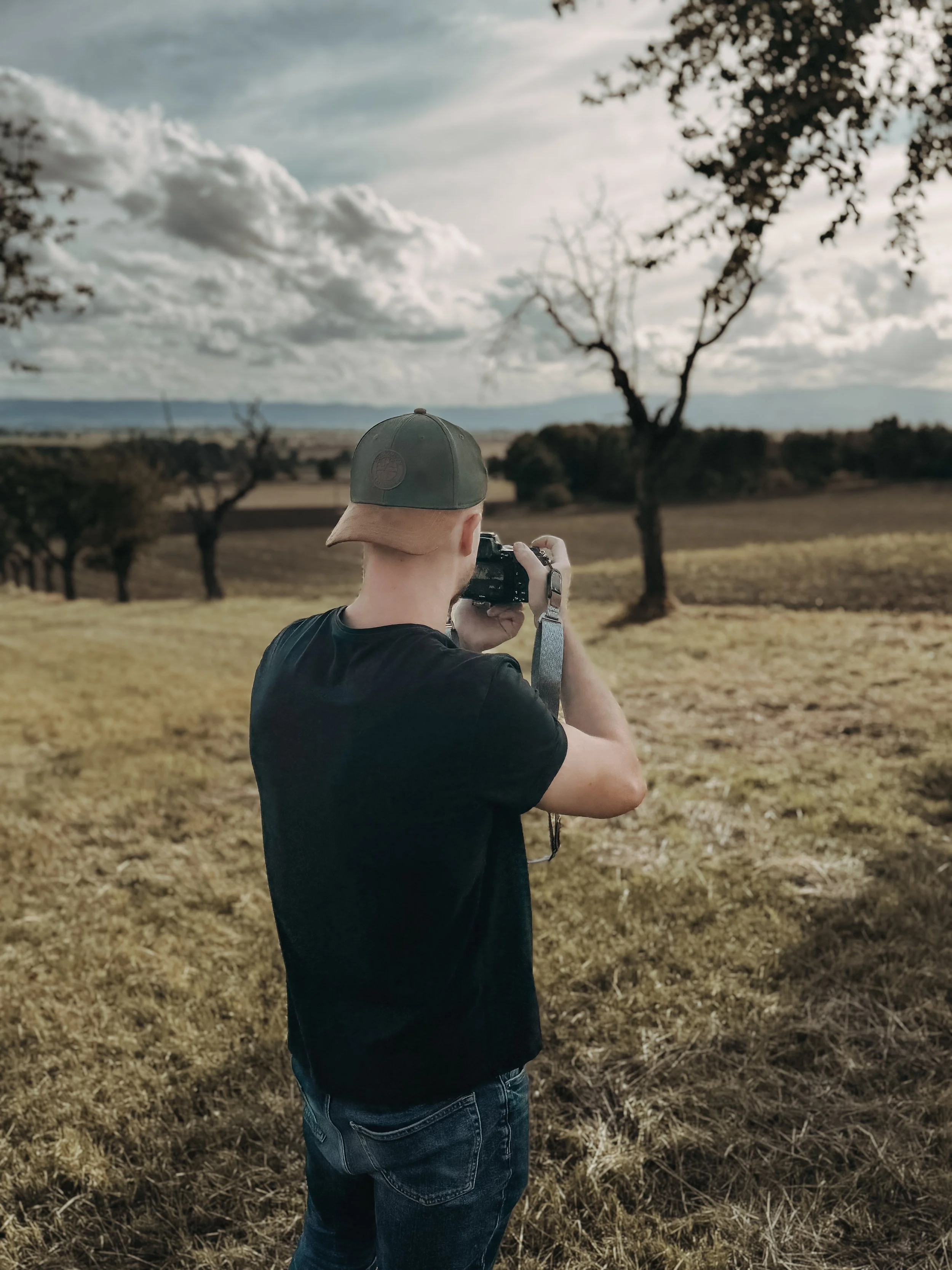Junge steht in einem offenen Feld, fotografiert die Landschaft mit einer Kamera, trägt eine grüne Cap und schwarze T-Shirt.