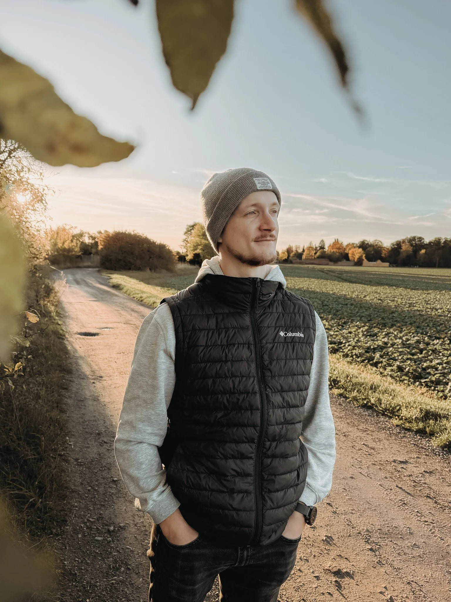 Ein junger Mann steht auf einem Feldweg bei Sonnenuntergang, trägt eine graue Mütze, schwarze Weste, graues Sweatshirt mit Hoodie, blickt in die Ferne.