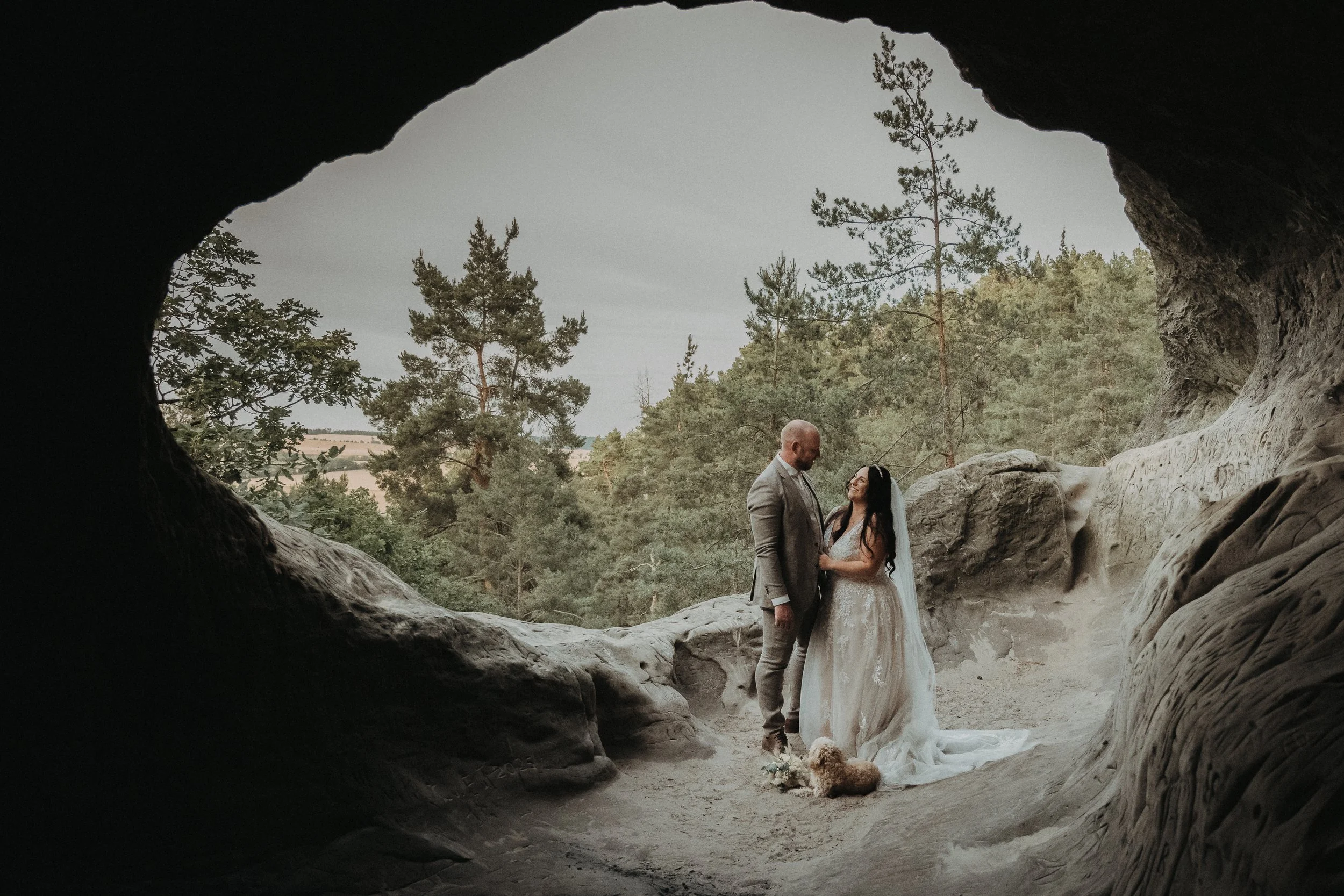Ein Brautpaar in Hochzeitskleidung in einer Felsennische, umgeben von Bäumen, mit Hund vor ihnen, in natürlicher Umgebung.