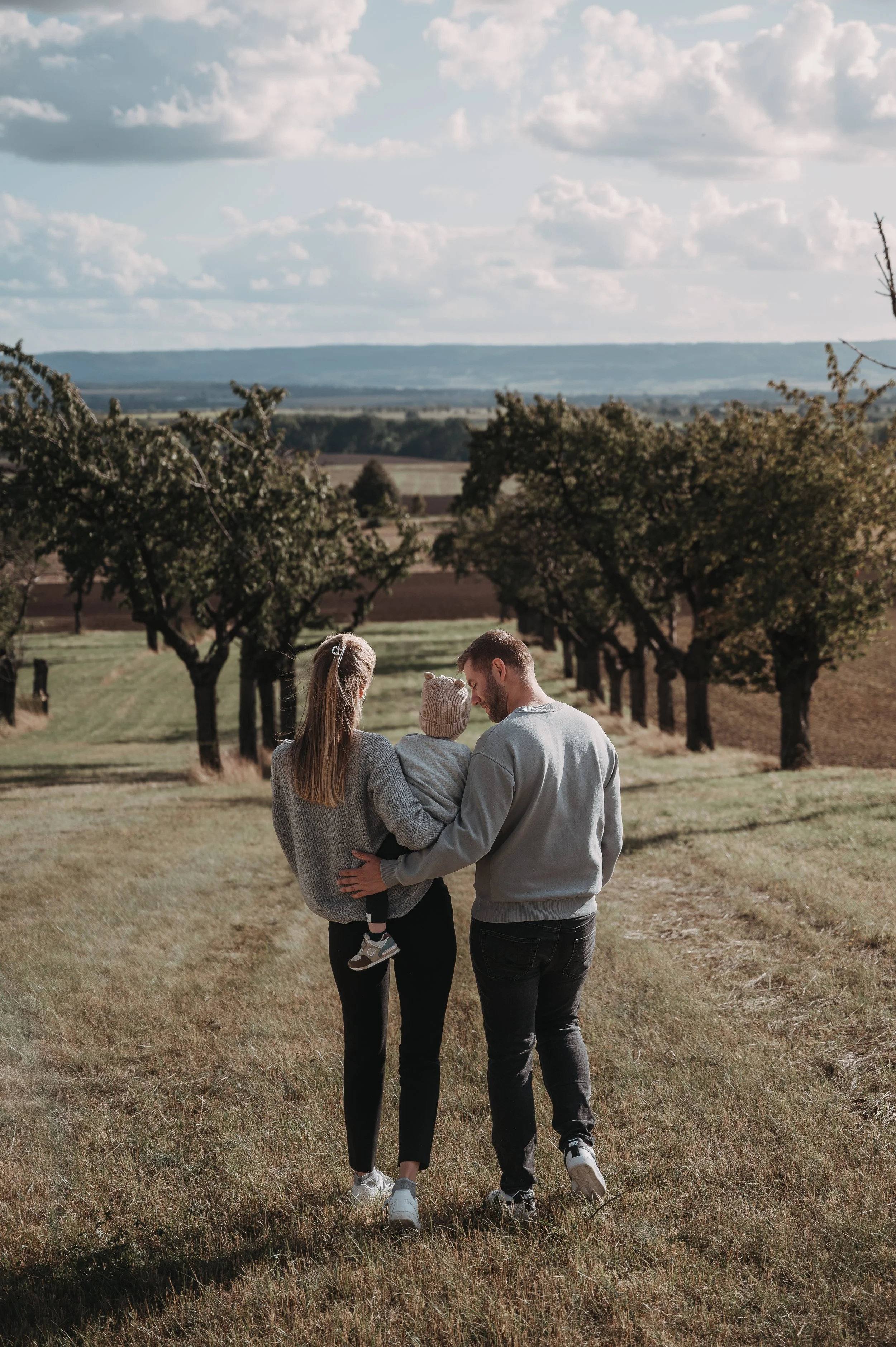 Familie mit Kind beim Spaziergang in der Natur, umliegende Bäume, weite offene Landschaft, leicht bewölkter Himmel.
