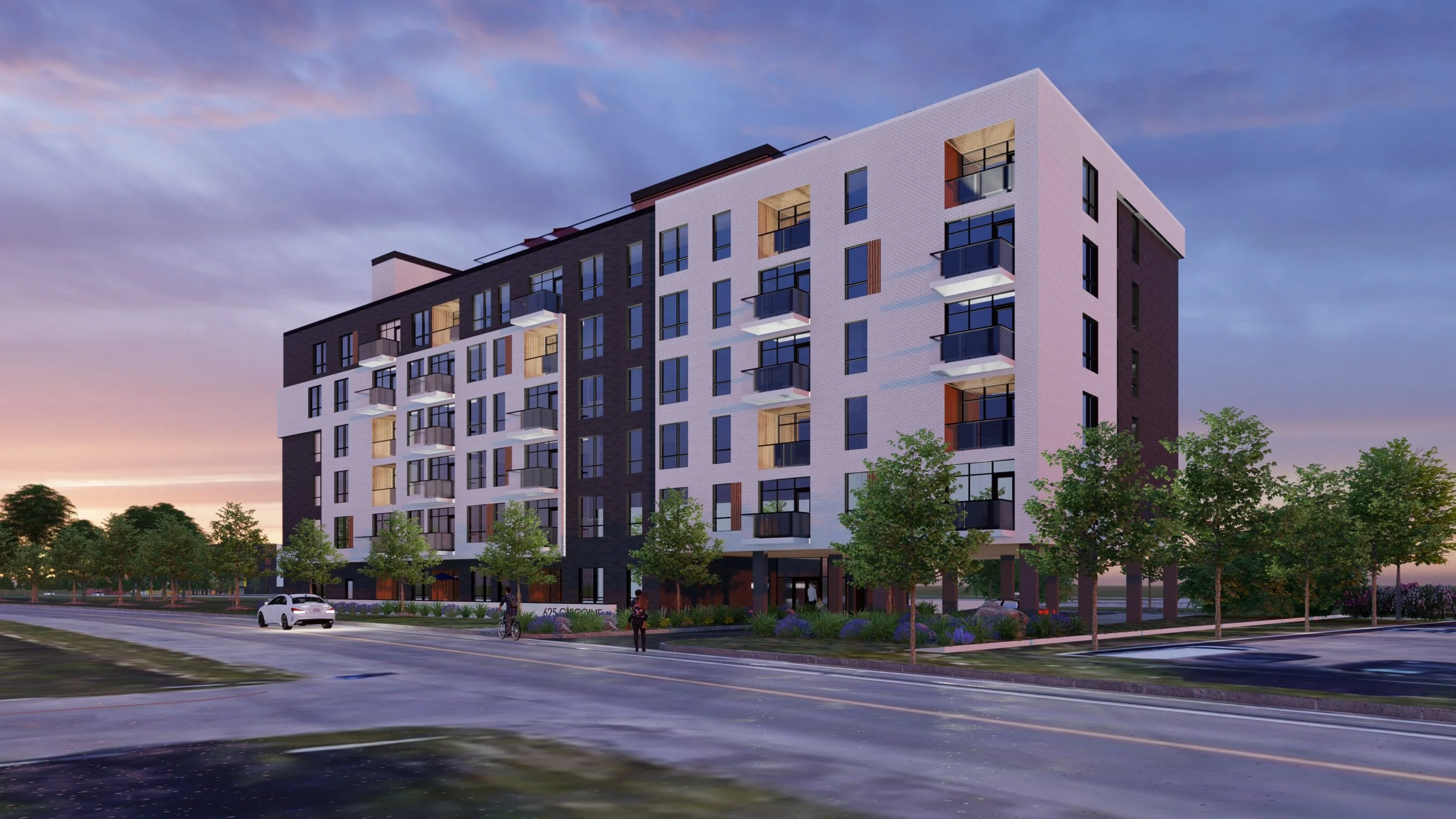 Un immeuble résidentiel moderne avec plusieurs balcons, entouré d'arbres et située près d'une route, avec un ciel au crépuscule.