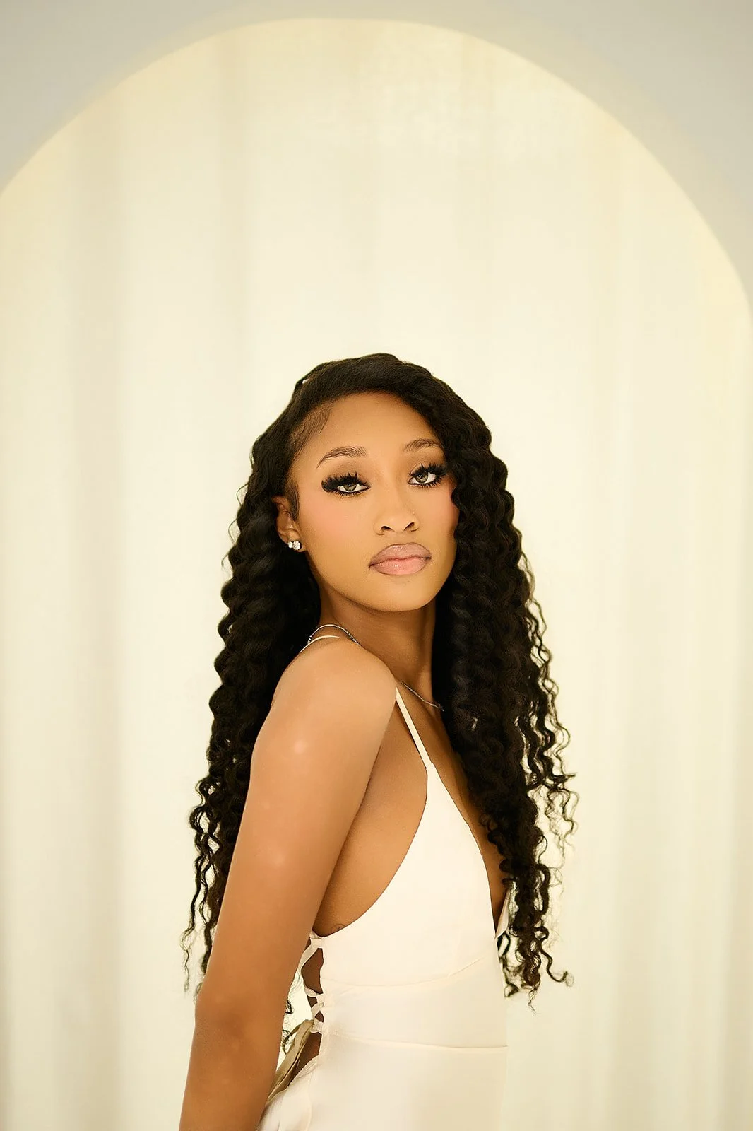 A woman with long curly black hair and makeup in a white dress, looking at the camera, standing in a softly lit room with an arched doorway in the background.