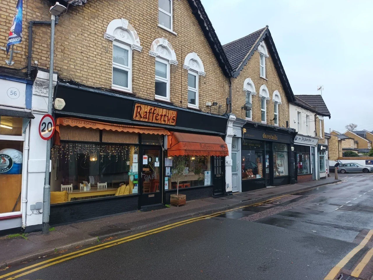 Street view of small shops with brick buildings, including a café called Rafferfy's with black and orange storefront, and nearby shops with different signage, on a wet day.