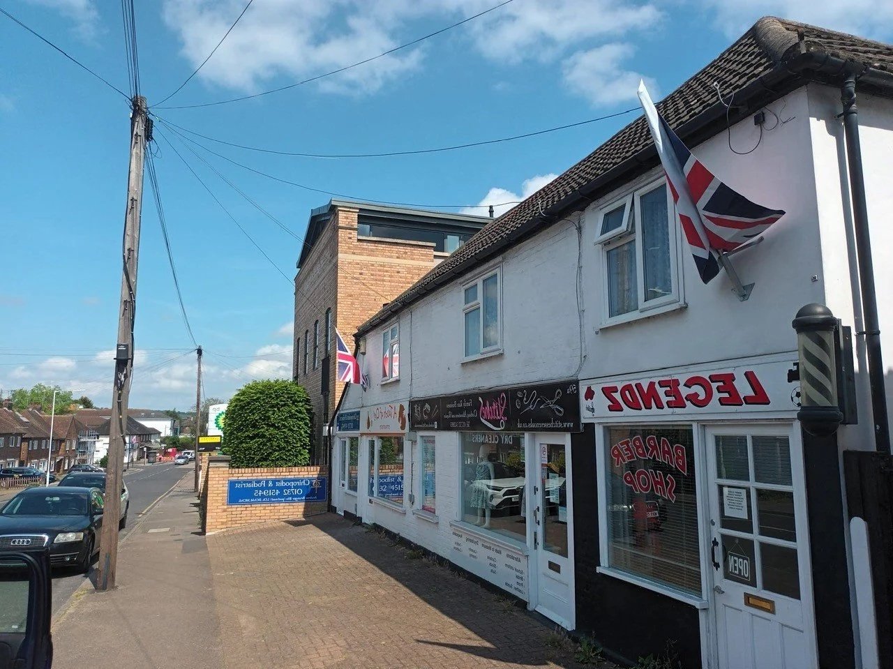 Street view of a British-themed shop with Union Jack flags, with signage reading 'Legend Barber Shop,' located on a sidewalk next to parked cars and residential buildings under a blue sky.