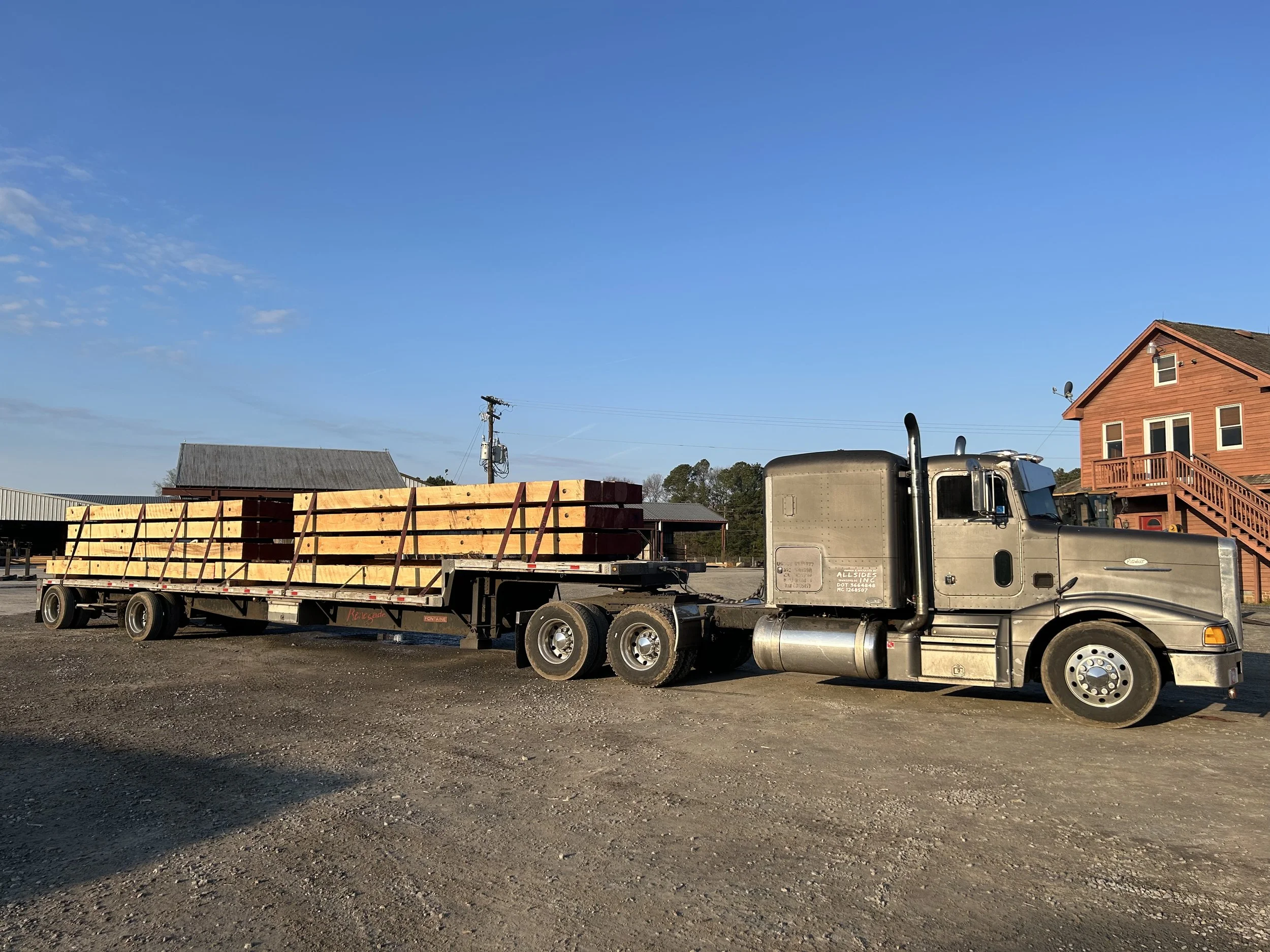 A semi-truck with a silver cab and trailer loaded with wooden planks parked on a gravel lot under a blue sky.