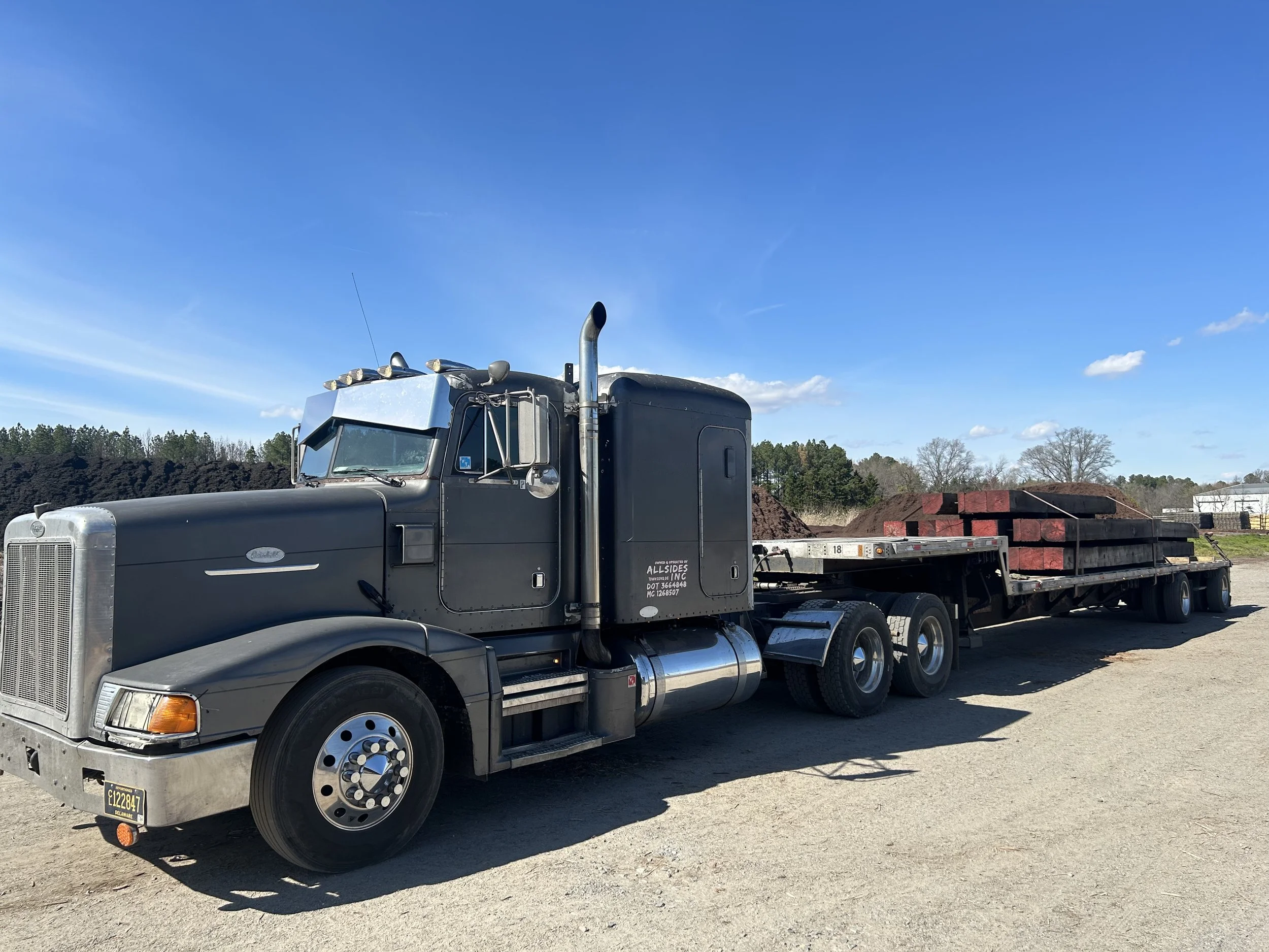 Black semi-truck parked on a gravel lot, carrying asphalt or similar material on the trailer, with a blue sky and some trees in the background.