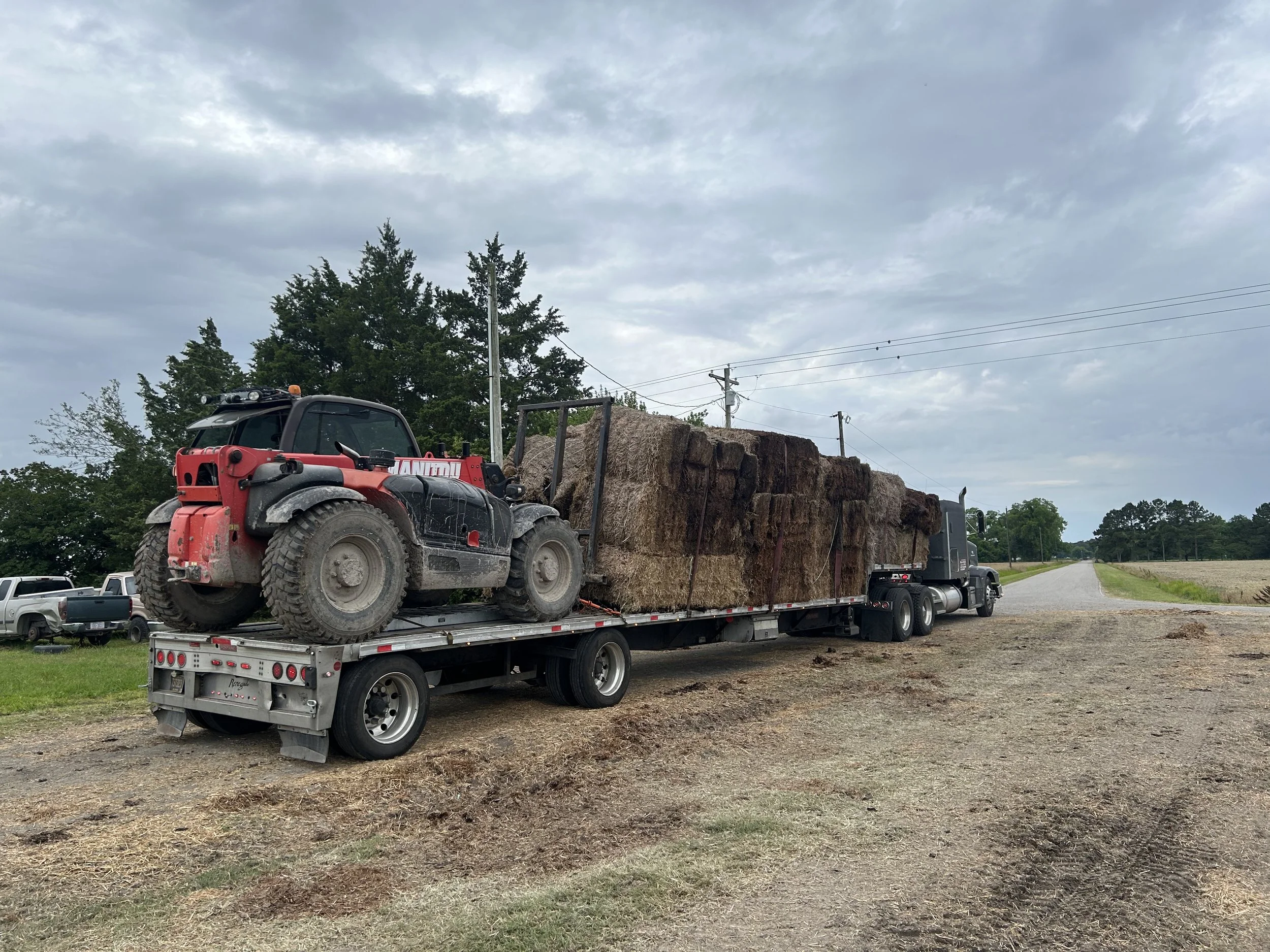 A semi-truck with a flatbed trailer carrying a load of hay bales parked on a rural roadside under cloudy skies.
