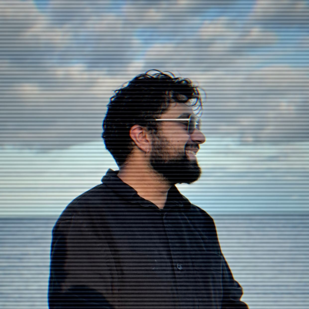 A man with a beard and curly hair smiling, wearing sunglasses and a black shirt, standing outdoors with a cloudy sky in the background.