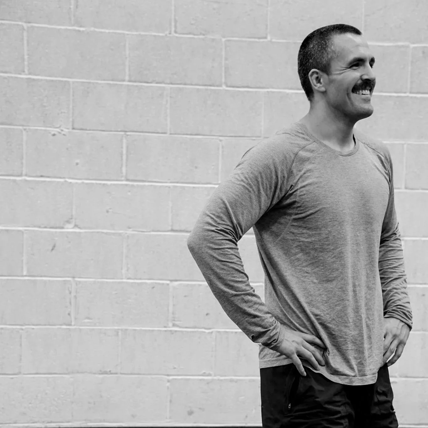 A smiling man standing with hands on his hips against a brick wall, wearing a long sleeve shirt and dark pants.