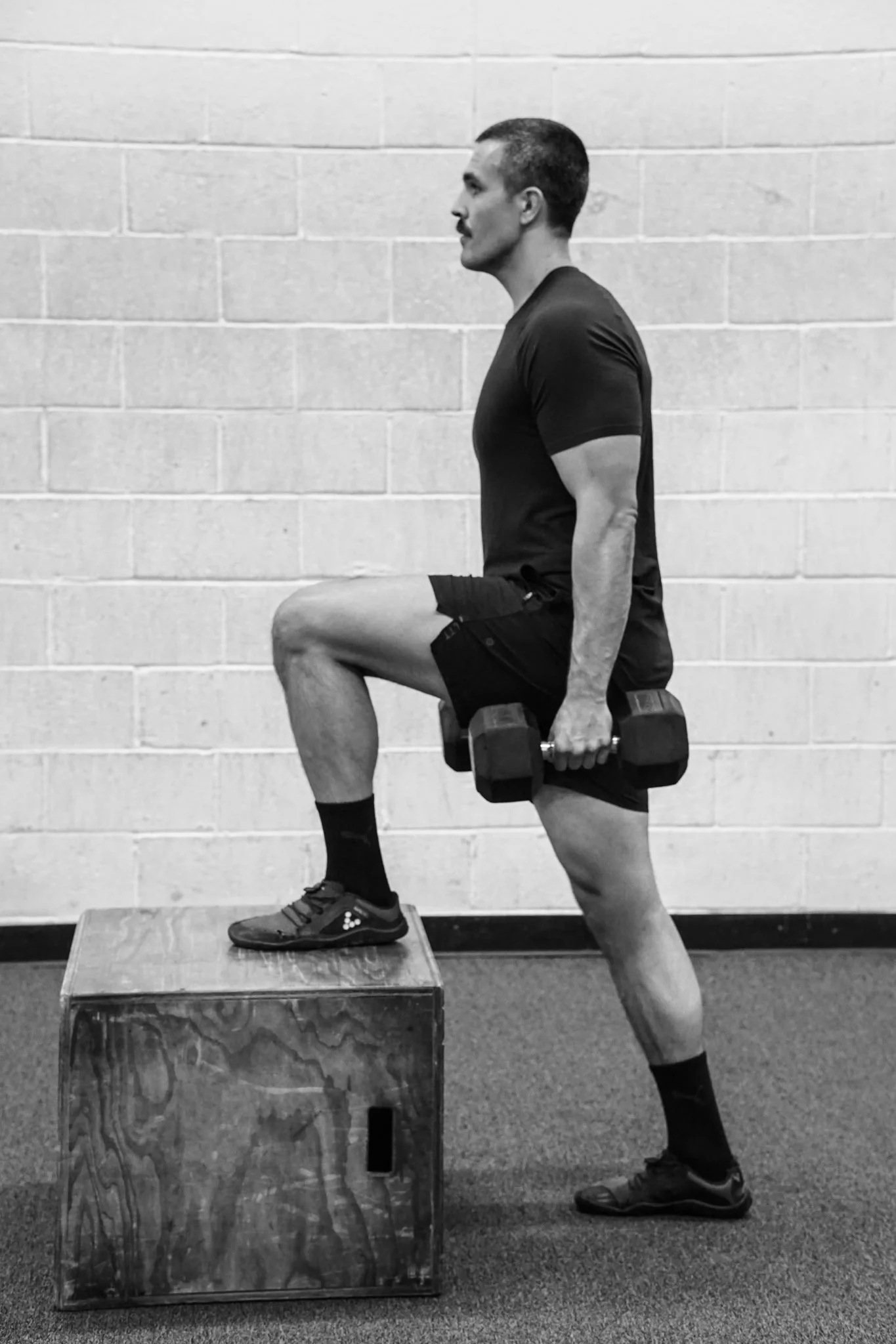 A man in athletic wear performing a step-up exercise with a dumbbell in each hand, standing on a wooden plyometric box in a gym.