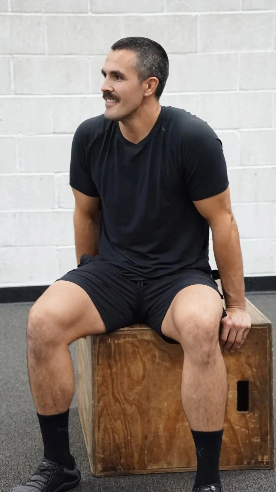A man sitting on a wooden plyo box in a gym, wearing a black t-shirt, black shorts, black socks, and black sneakers, with a gray brick wall background.