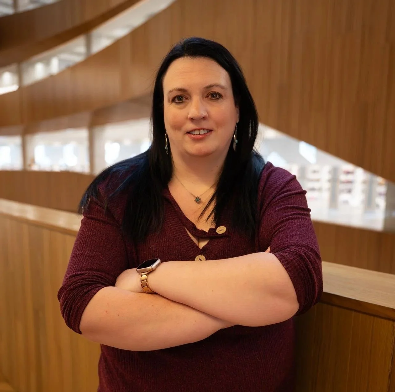 A woman with black hair and light skin, wearing a maroon sweater, standing with arms crossed in an indoor setting with wooden wall panels and large windows.