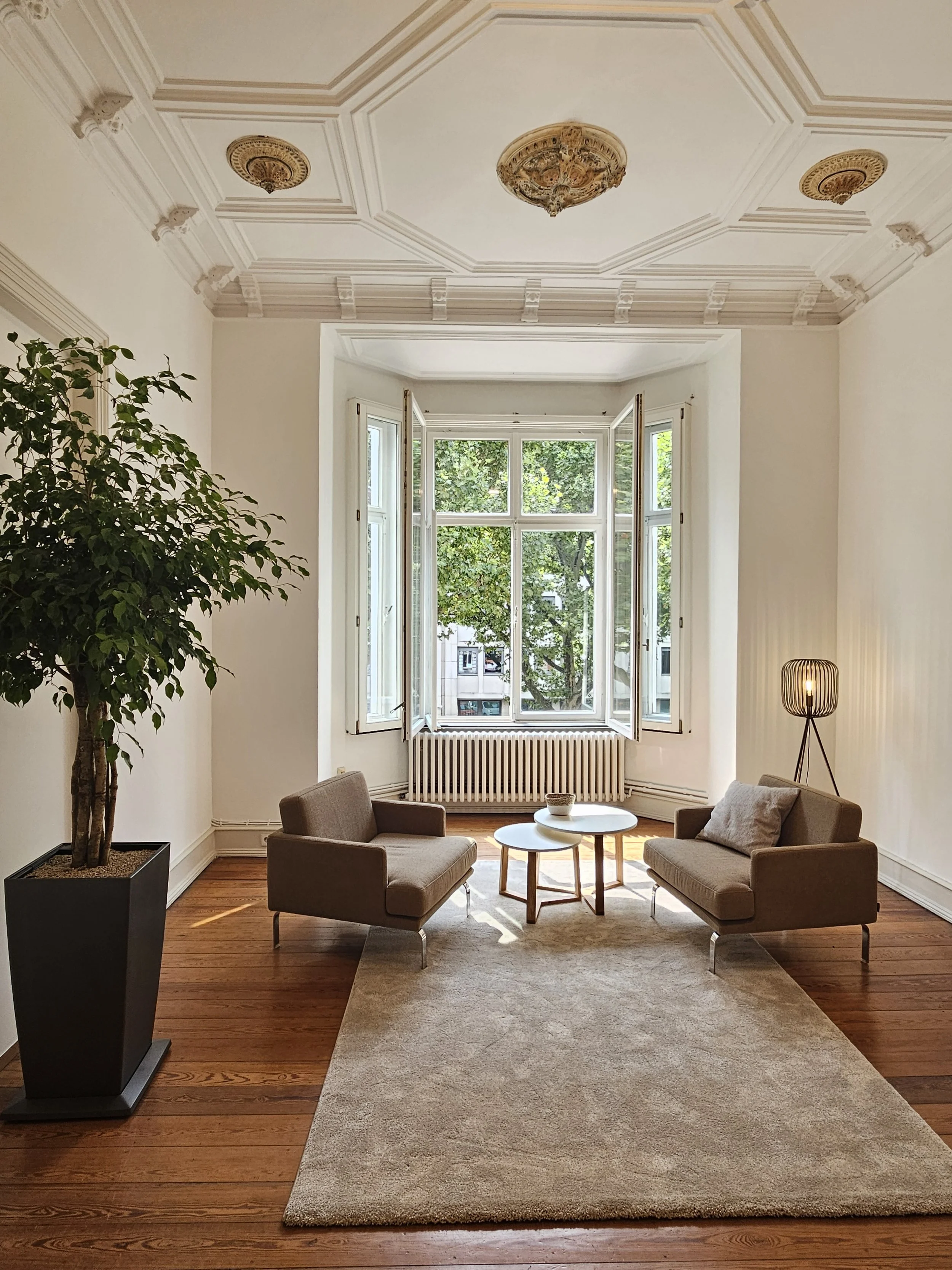 Ein Wohnzimmer mit weiß gestrichenen Wänden, einer großen Fensterfront, einem beige Teppich, zwei grauen Sofas mit Kissen, einem runden Couchtisch, einer Stehlampe und einer Topfpflanze.