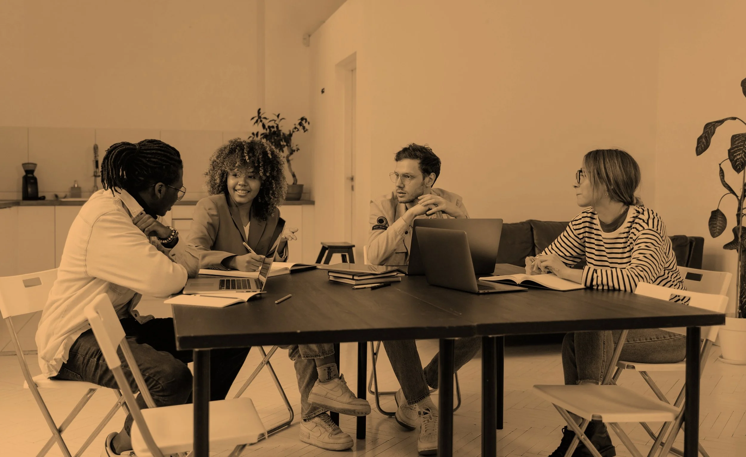 Four people having a meeting around a table with laptops, notebooks, and pens in a room with minimalistic decor and a potted plant in the background.