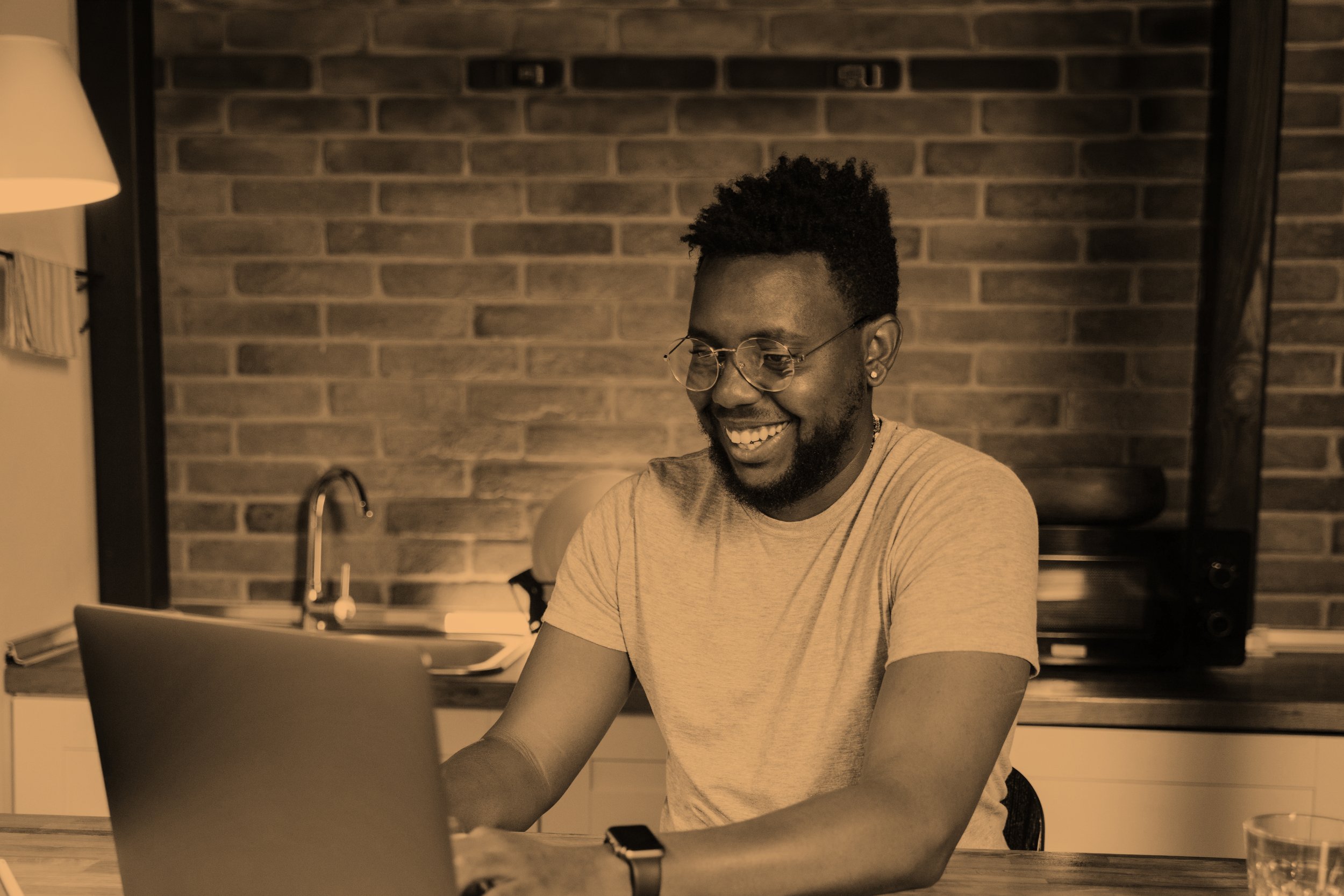 A smiling man with glasses and a beard working on a laptop in a kitchen with exposed brick wall.