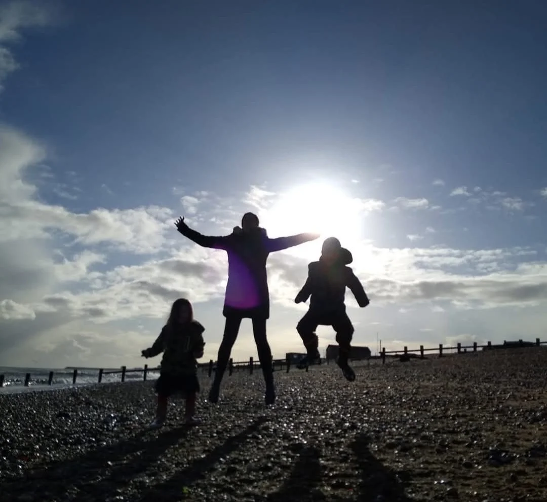 Children playing and jumping on a pebble beach with a fence, with the sun setting or rising in the background and clouds in the sky.