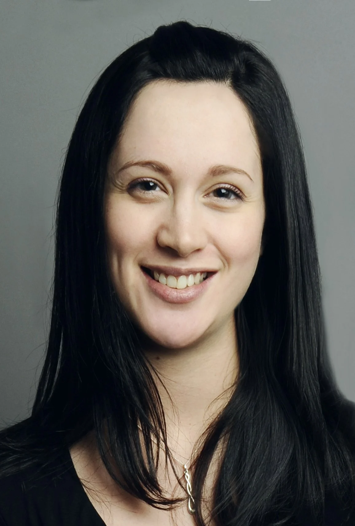 Headshot of a woman with long dark hair, smiling, wearing a black top and a silver necklace, against a gray background.