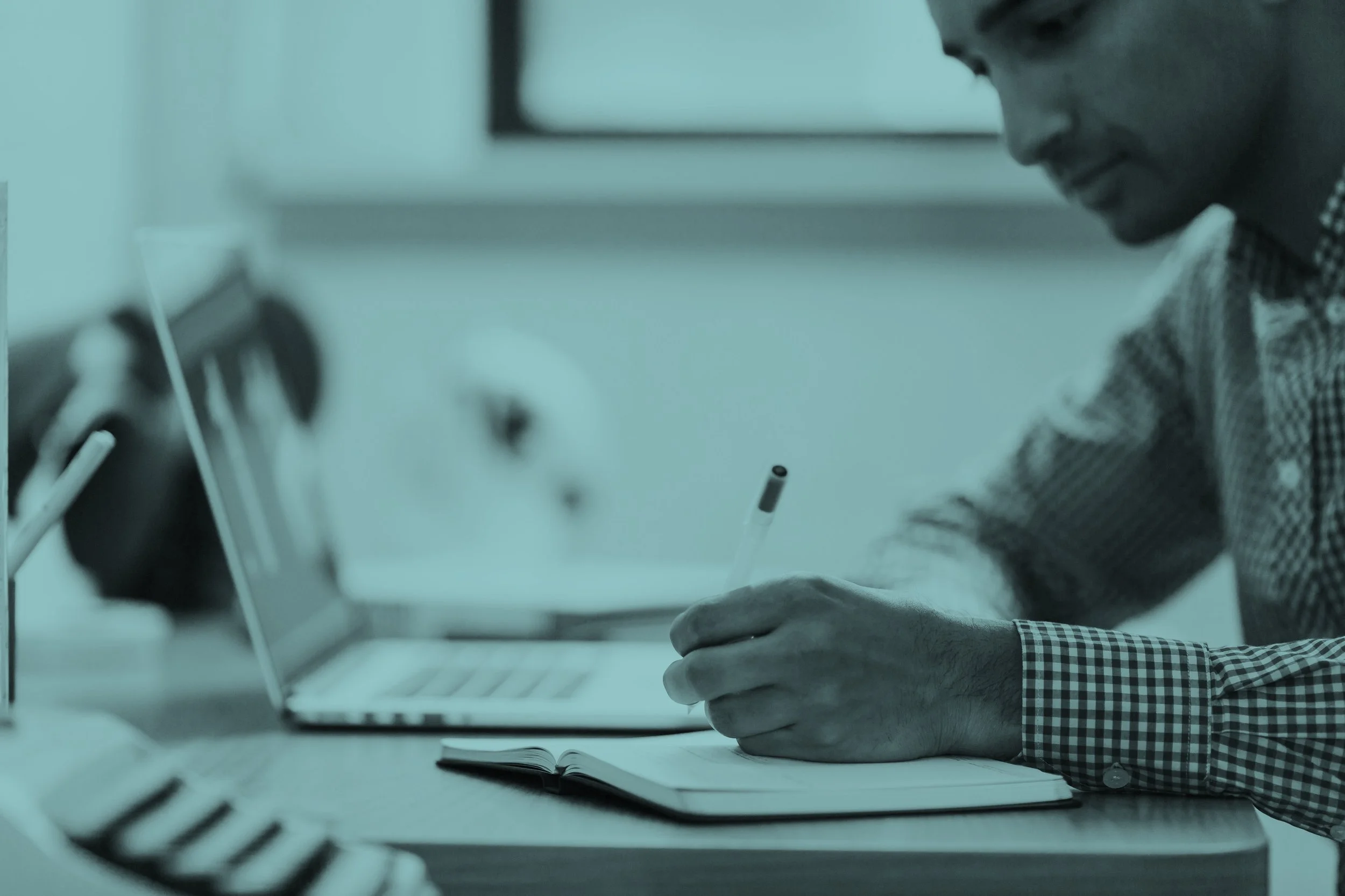 A man sitting at a desk, writing in a notebook with a pen, with a laptop and other office supplies on the desk.