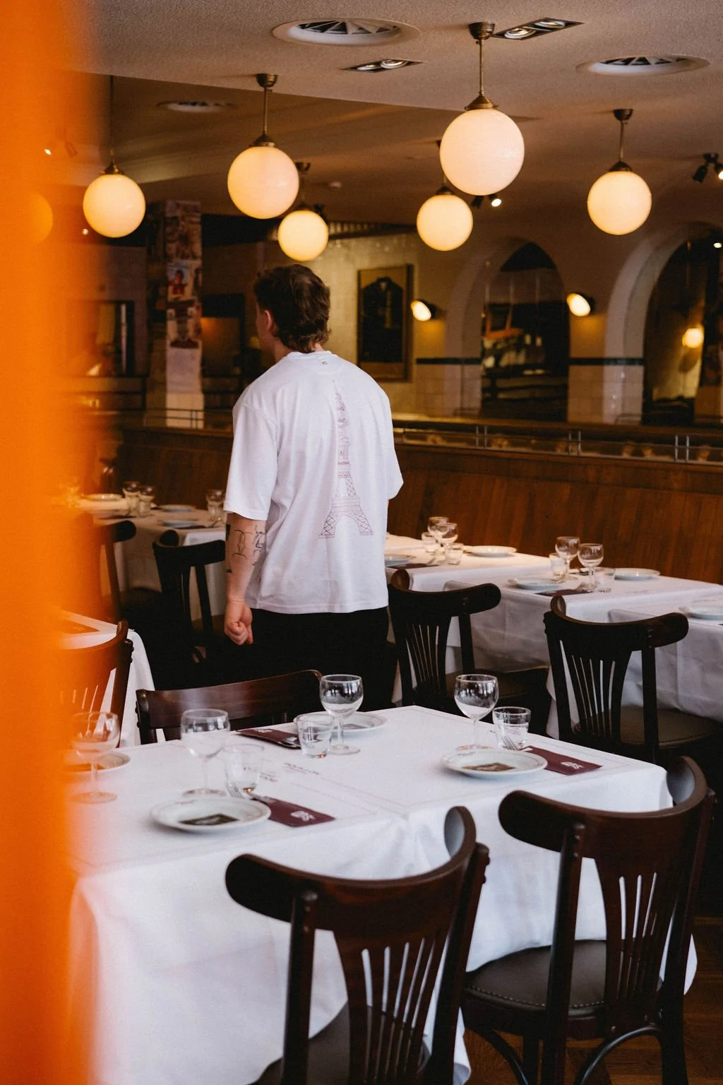 A person standing in a restaurant with white-tablecloth-covered tables set with glasses and plates, hanging spherical pendant lights overhead, and a warm ambient lighting.